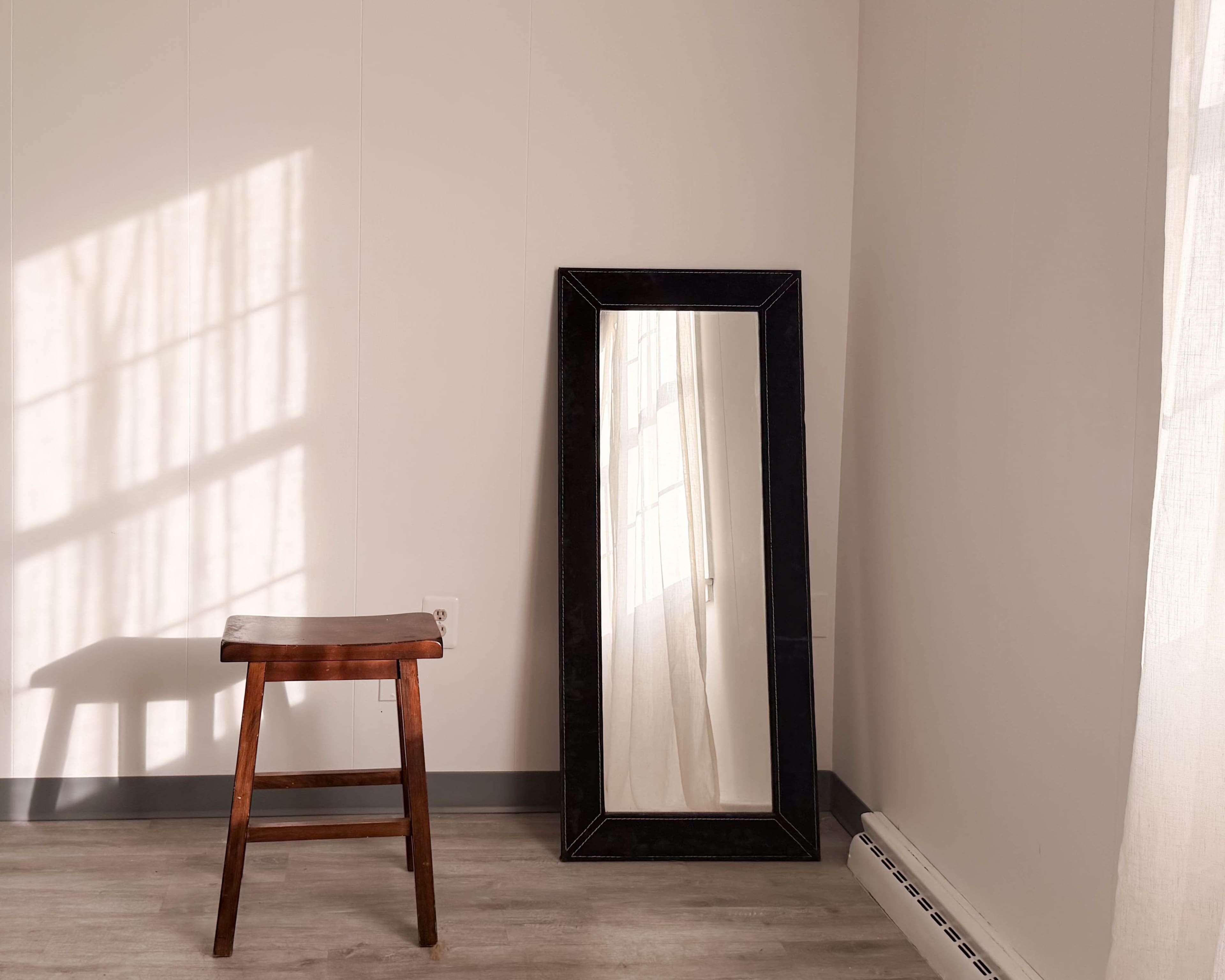 A wooden stool stands beside a tall mirror reflecting sunlight through a nearby window onto a light-colored wall.