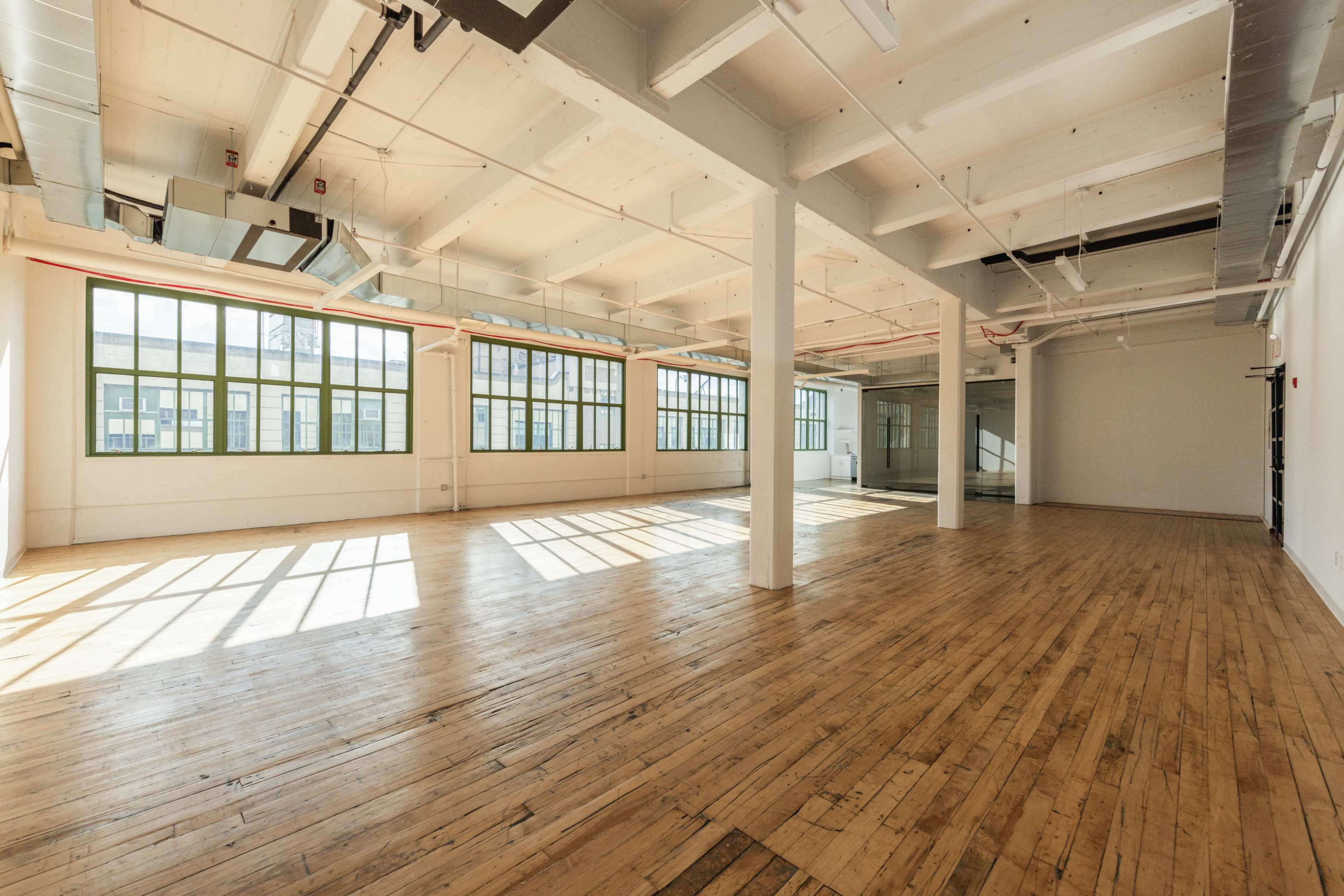 The image shows a large, empty interior space featuring wooden floors, white walls, and numerous windows allowing natural light to fill the room.