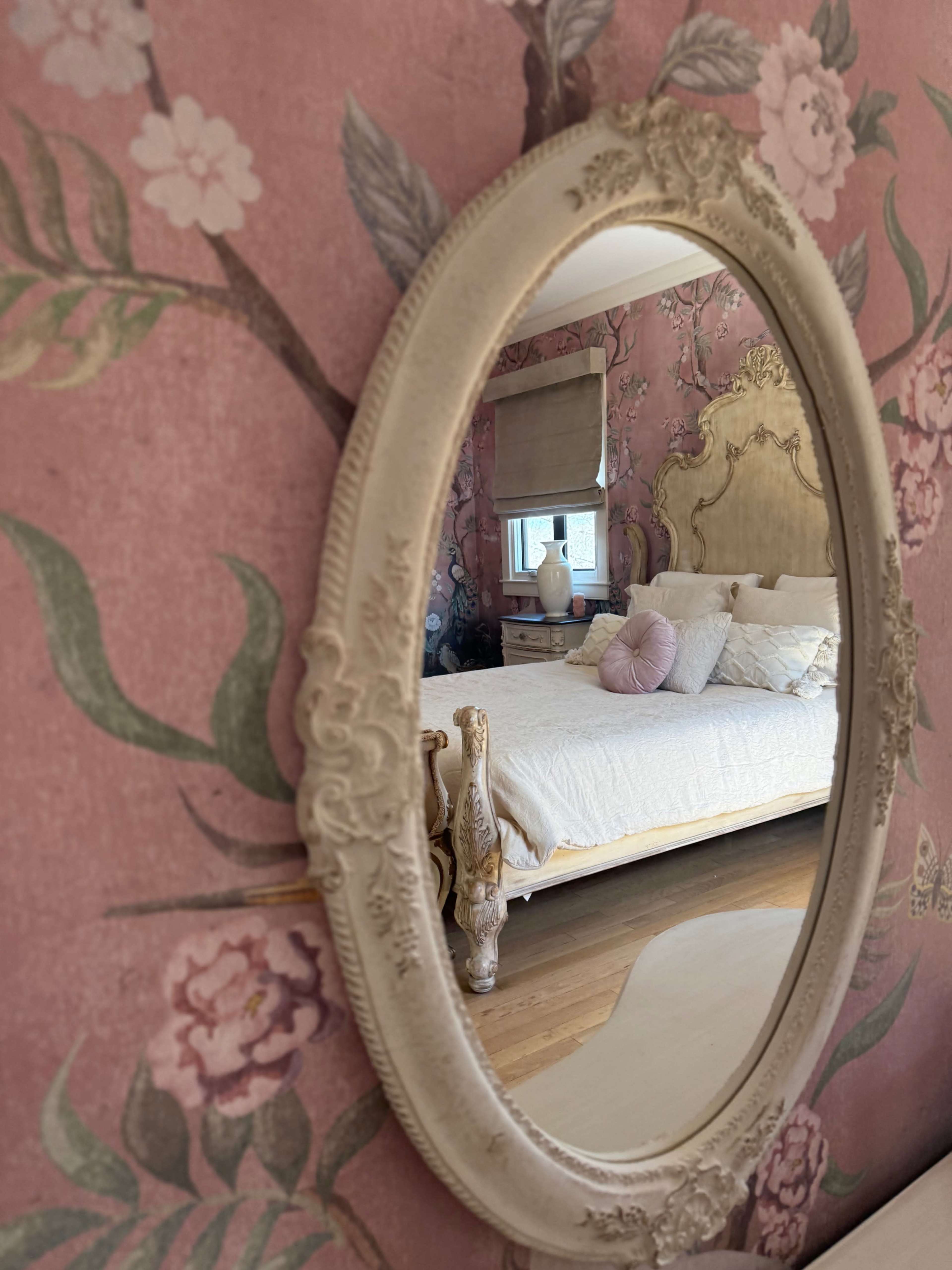 The image shows an ornate mirror reflecting a decorated bedroom with floral wallpaper and a neatly made bed.