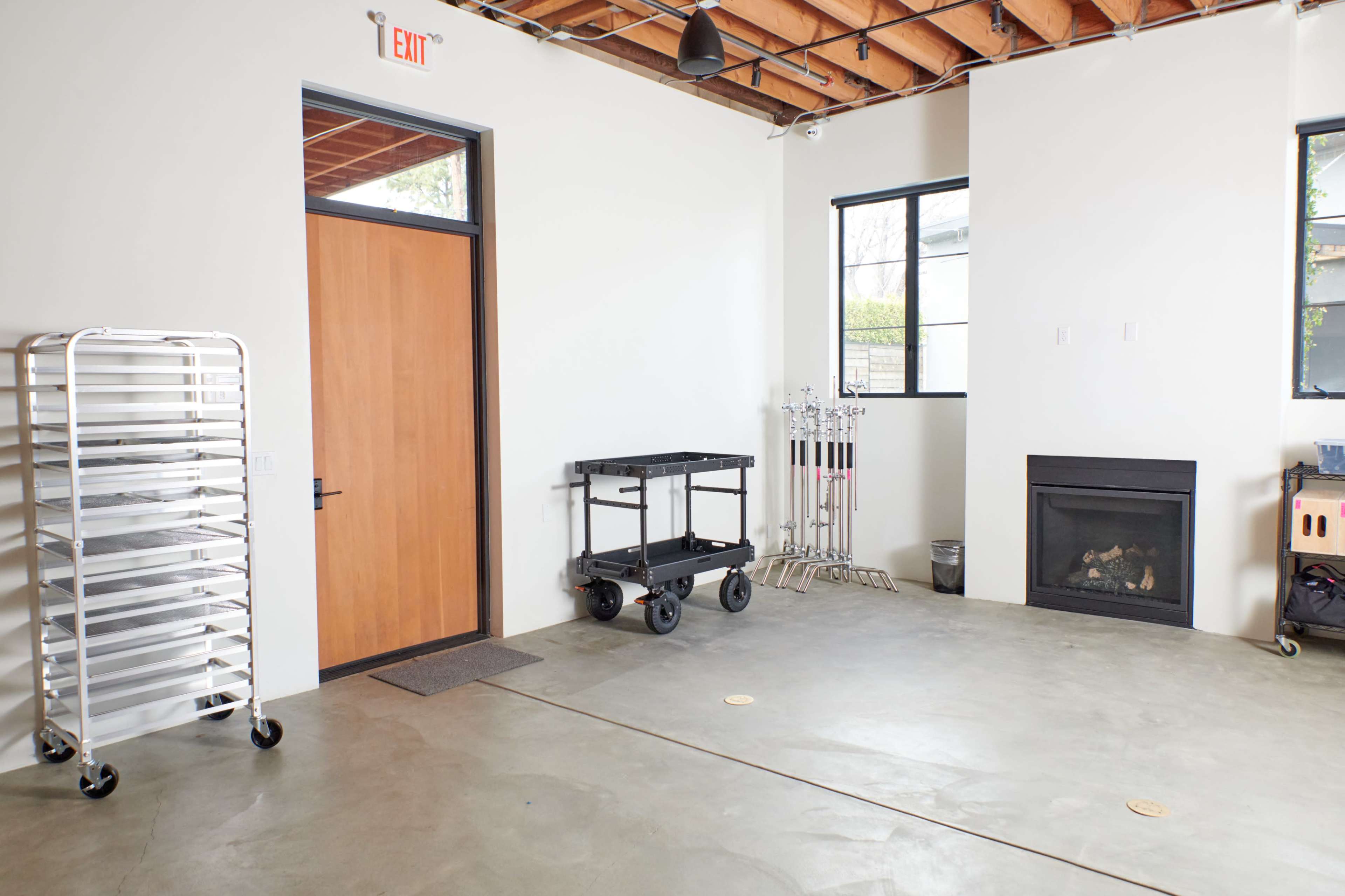The image shows a spacious room with a concrete floor, a wooden door, a black cart, a fireplace, and a metal rack against the walls.