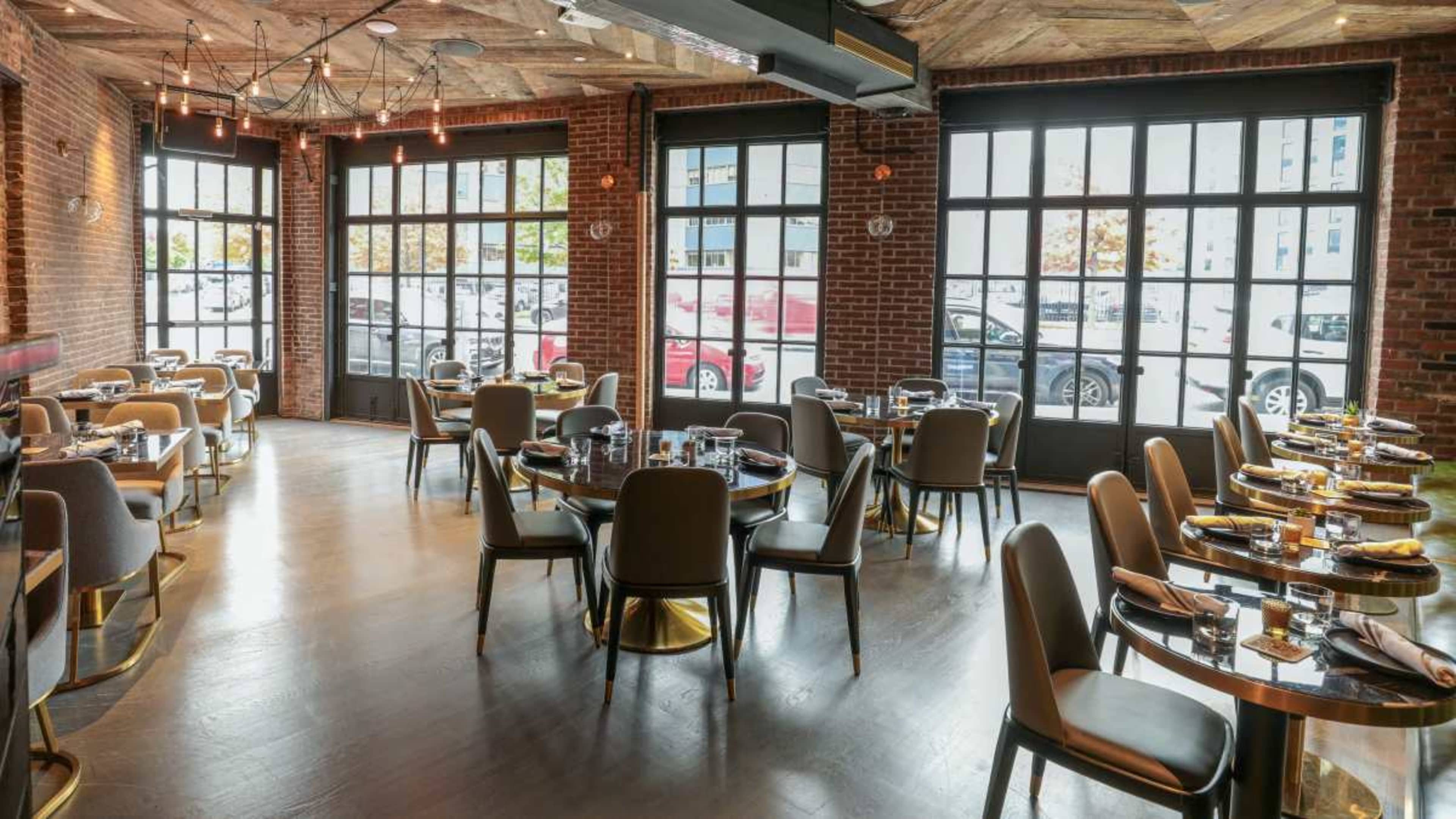 The image shows a modern restaurant interior with round tables set for dining, featuring exposed brick walls and large windows.