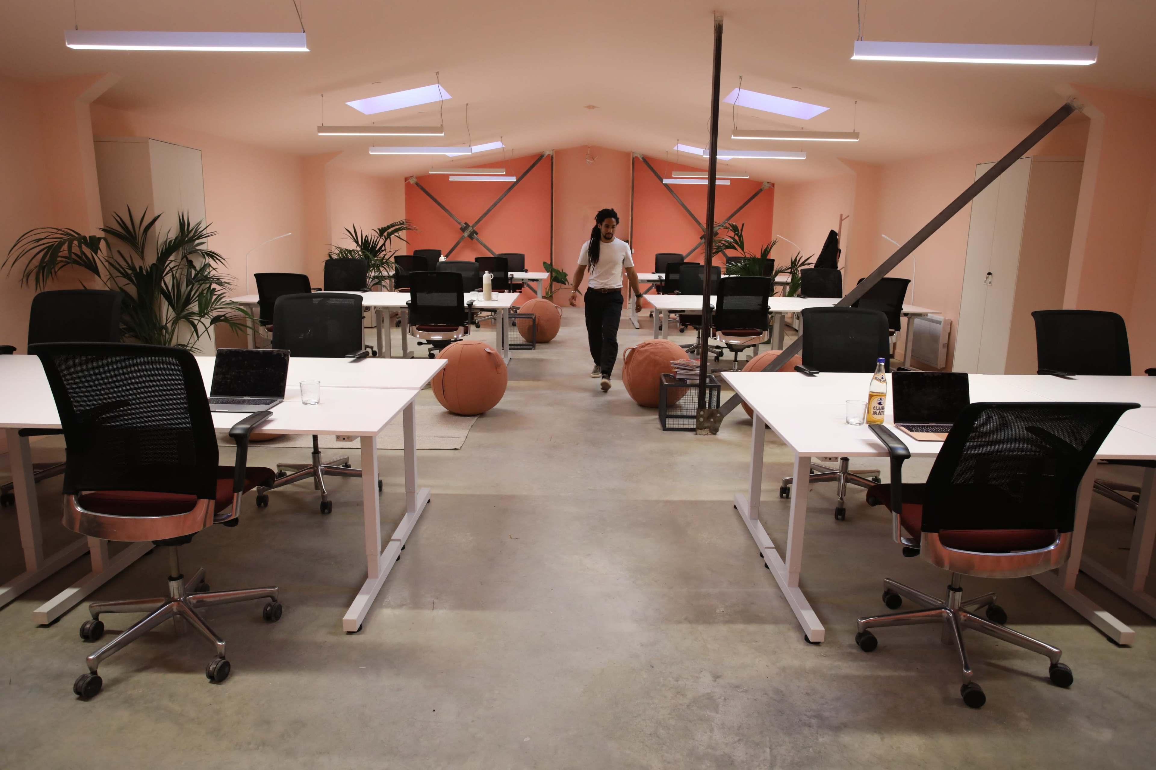 A woman walks through a spacious office with several desks, chairs, and plants under soft lighting.