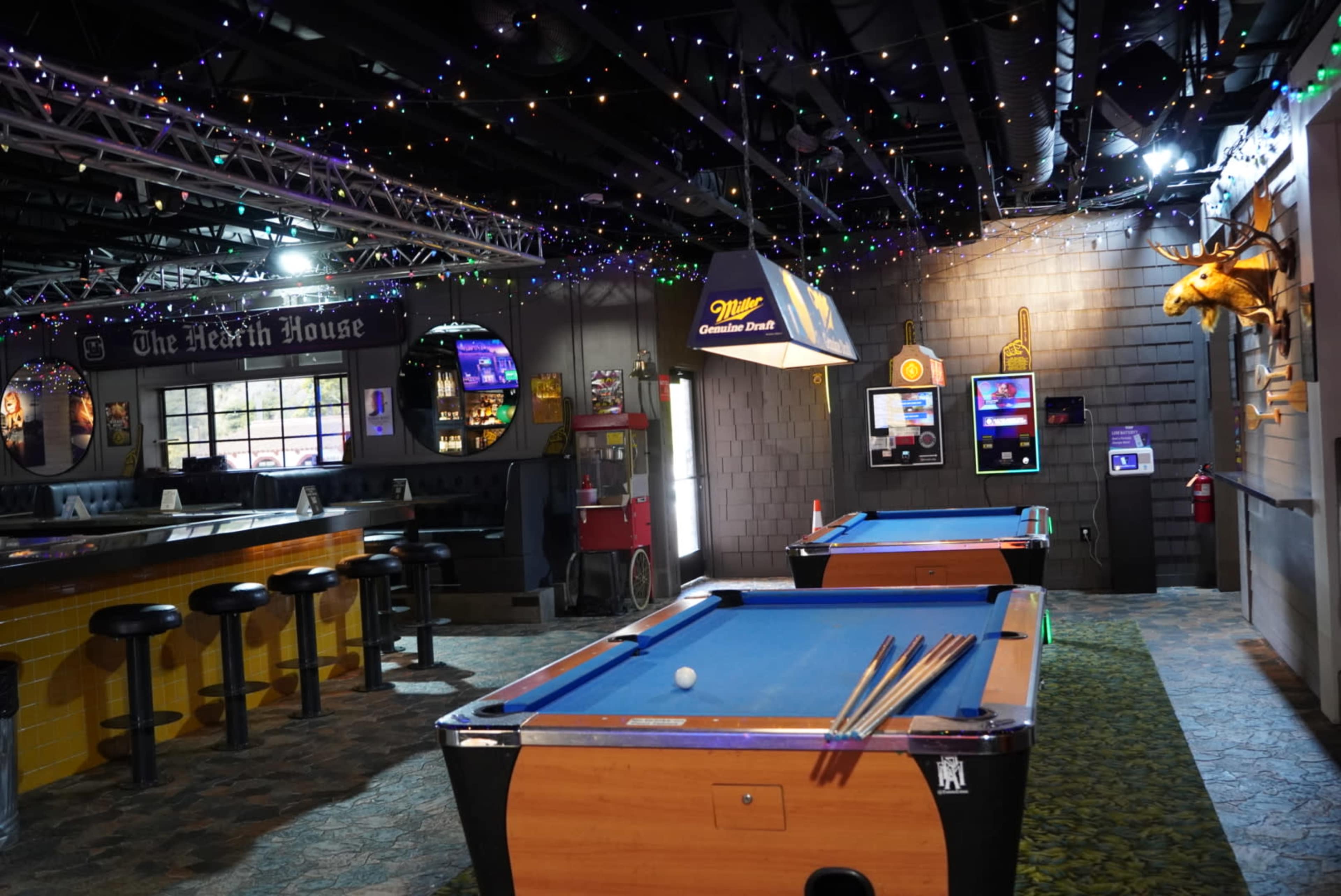 The image shows a dimly lit bar area featuring two pool tables, a long counter with bar stools, and decorative lights hanging from the ceiling.