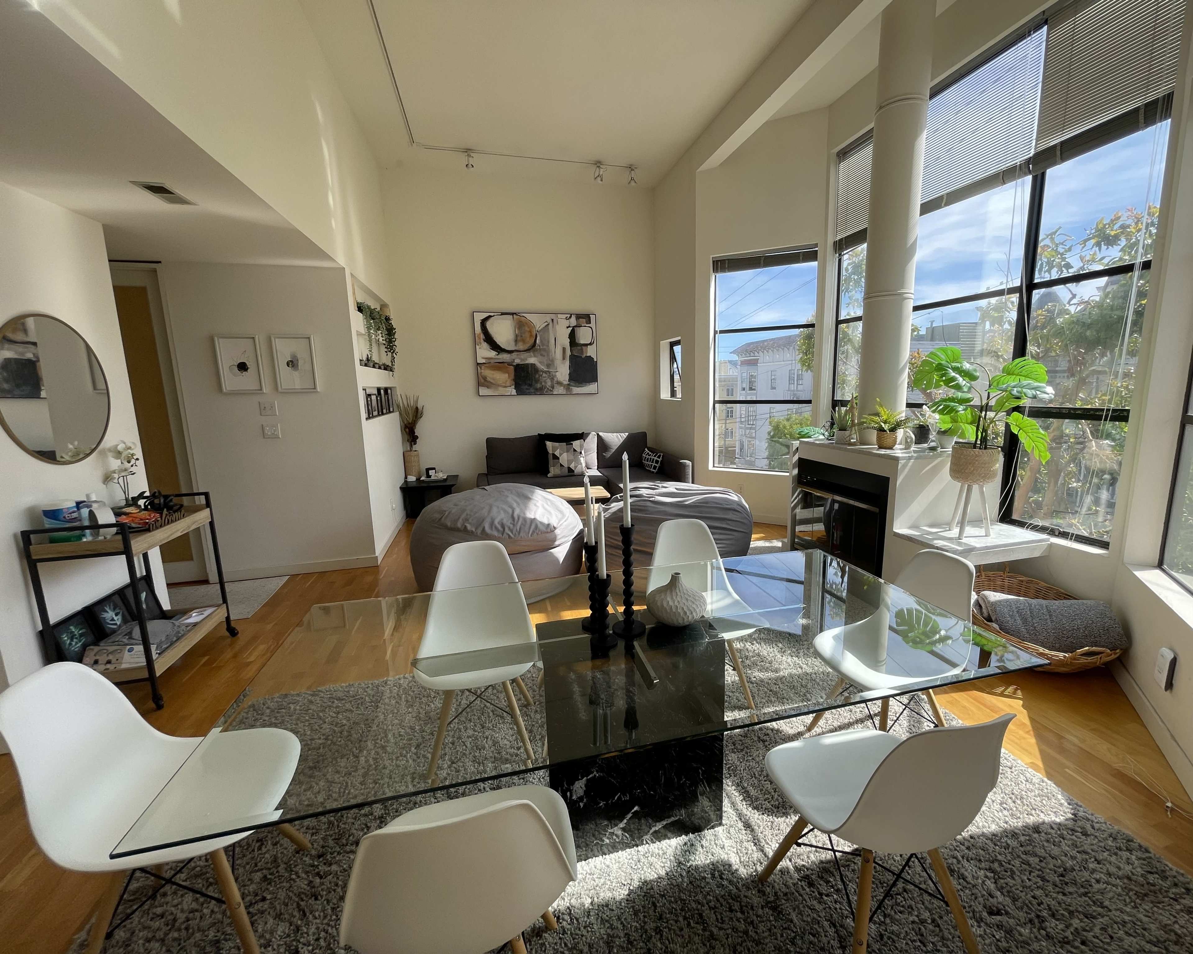 The image shows a modern dining area with a glass table surrounded by white chairs, adjacent to a spacious living area featuring a bed and large windows.