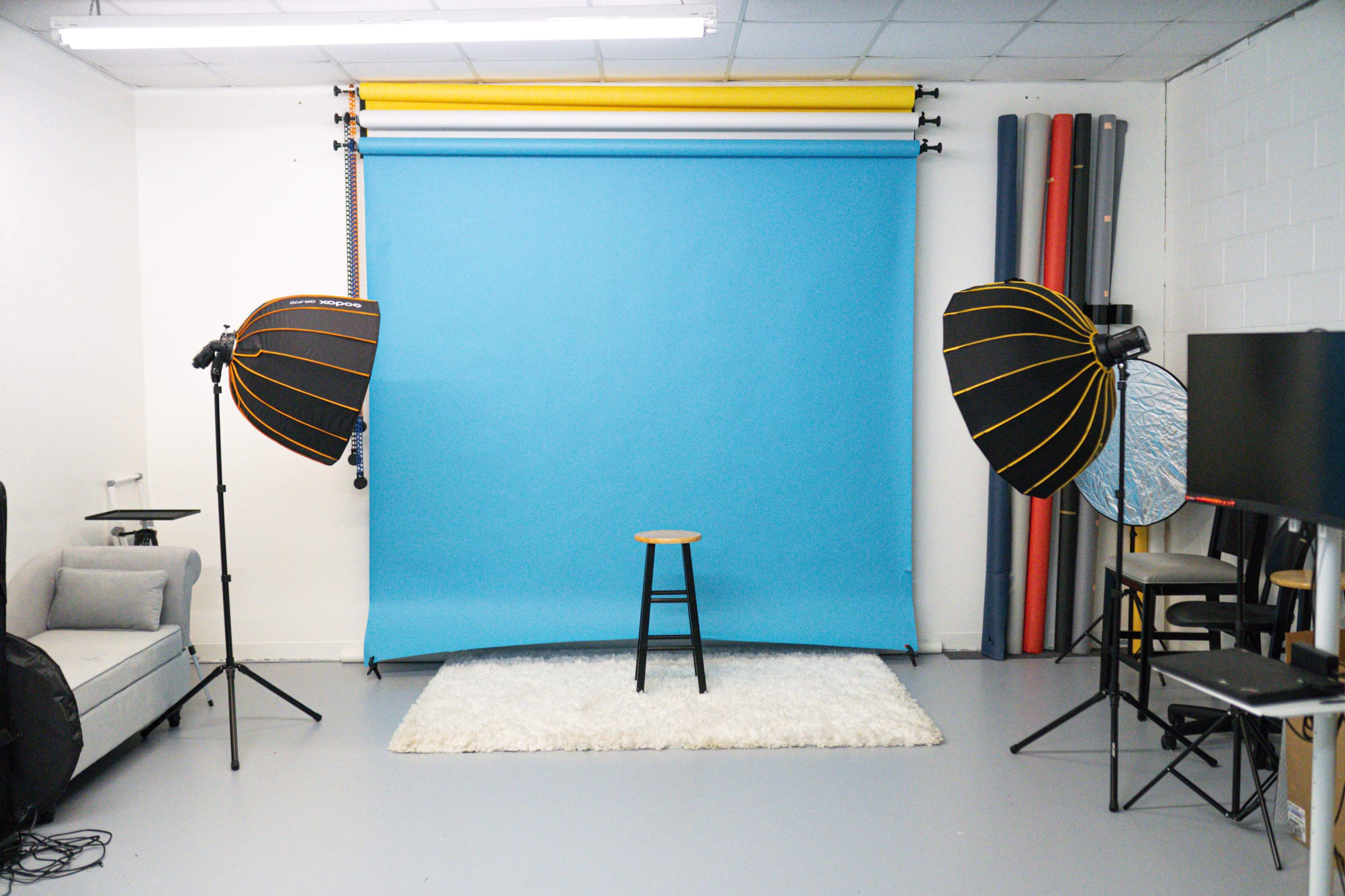 A photography studio equipped with a blue backdrop, two softbox lights, a stool, and a rug.
