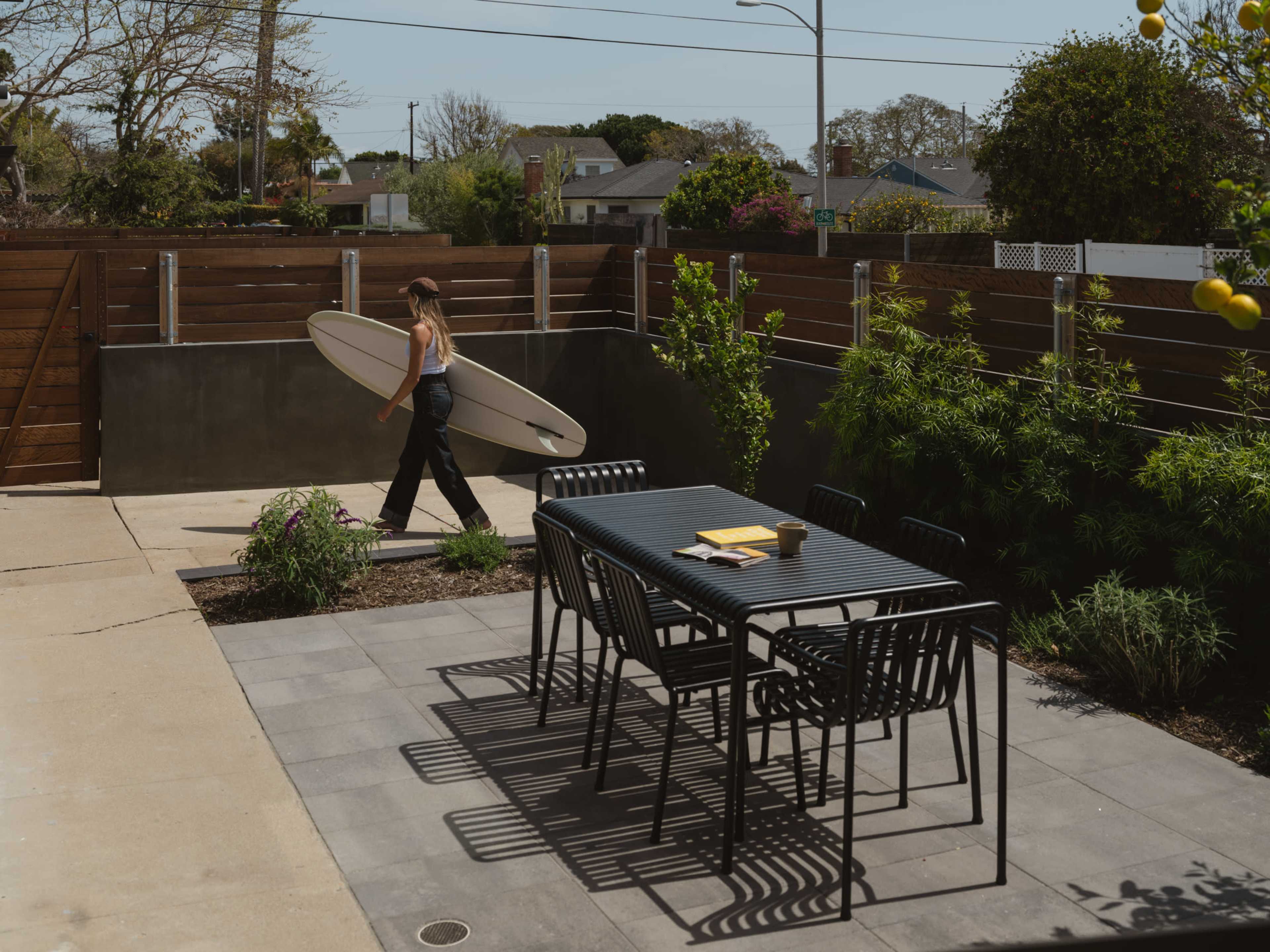 A person carrying a surfboard walks through a paved outdoor area, which features a dining table and lush greenery.