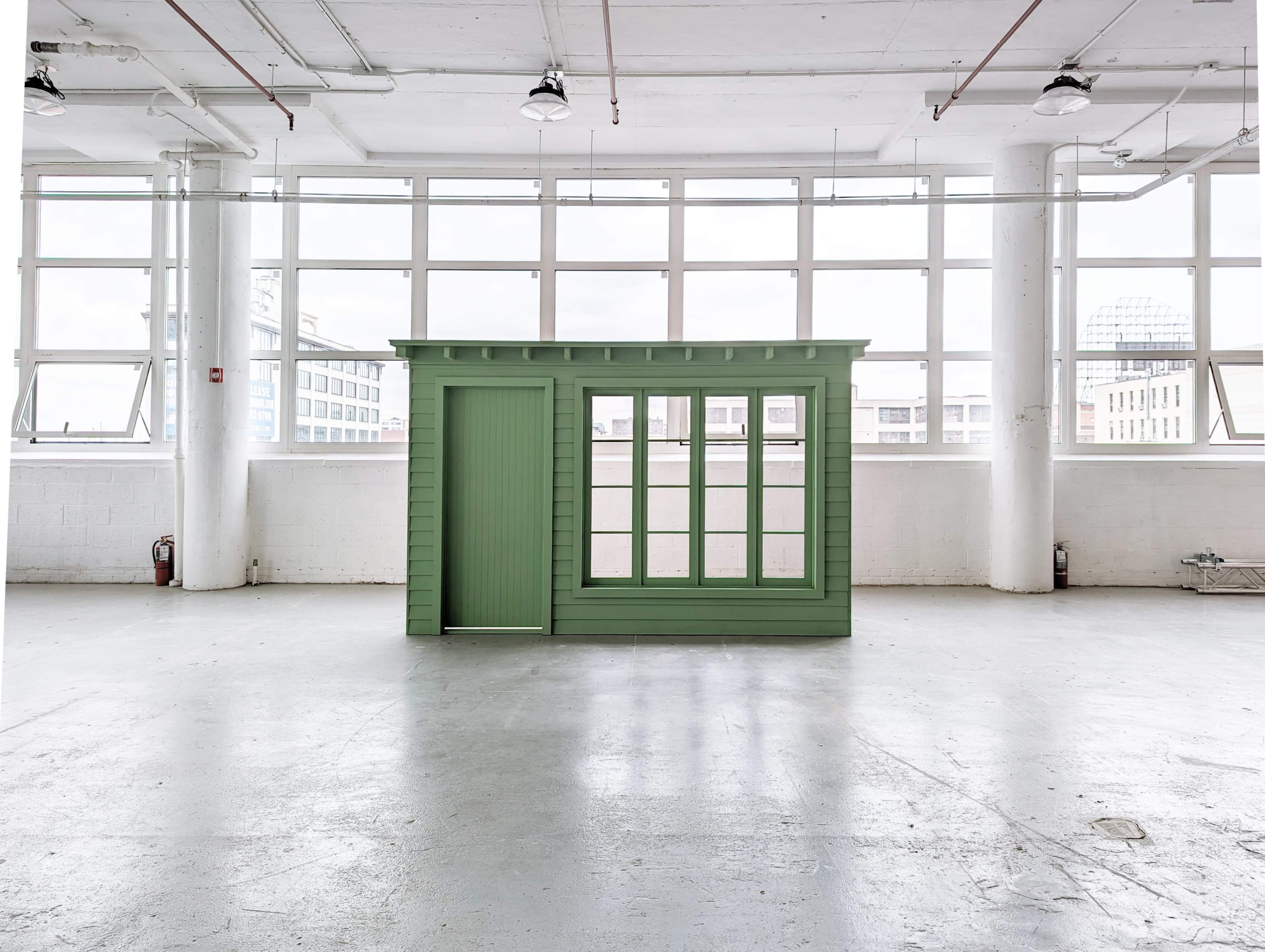 A green structure resembling a small building or shed positioned in an empty, well-lit industrial space with large windows.