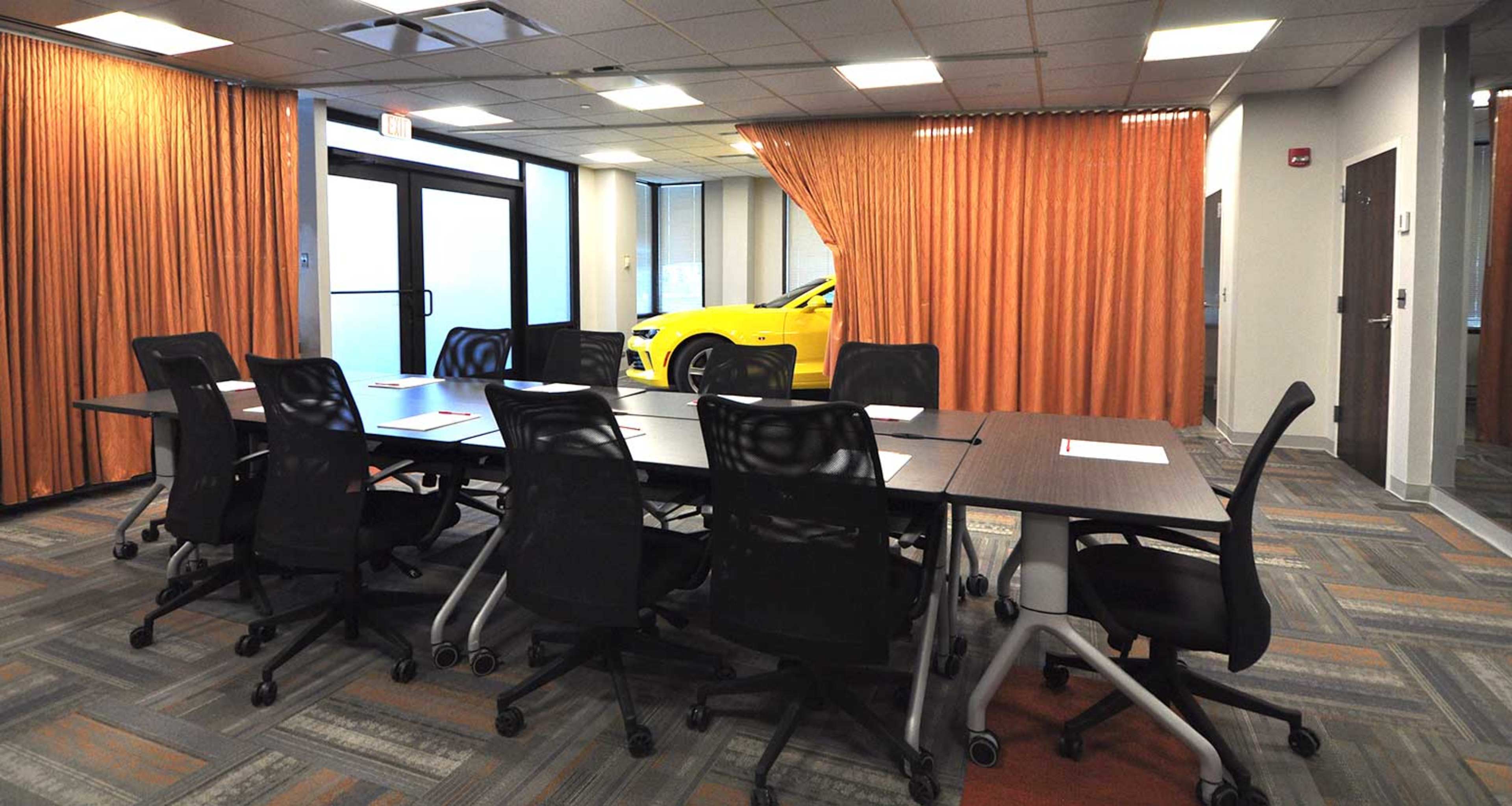 A conference room features a large table surrounded by black chairs, with sheer orange curtains partially drawn to reveal a yellow car parked outside.