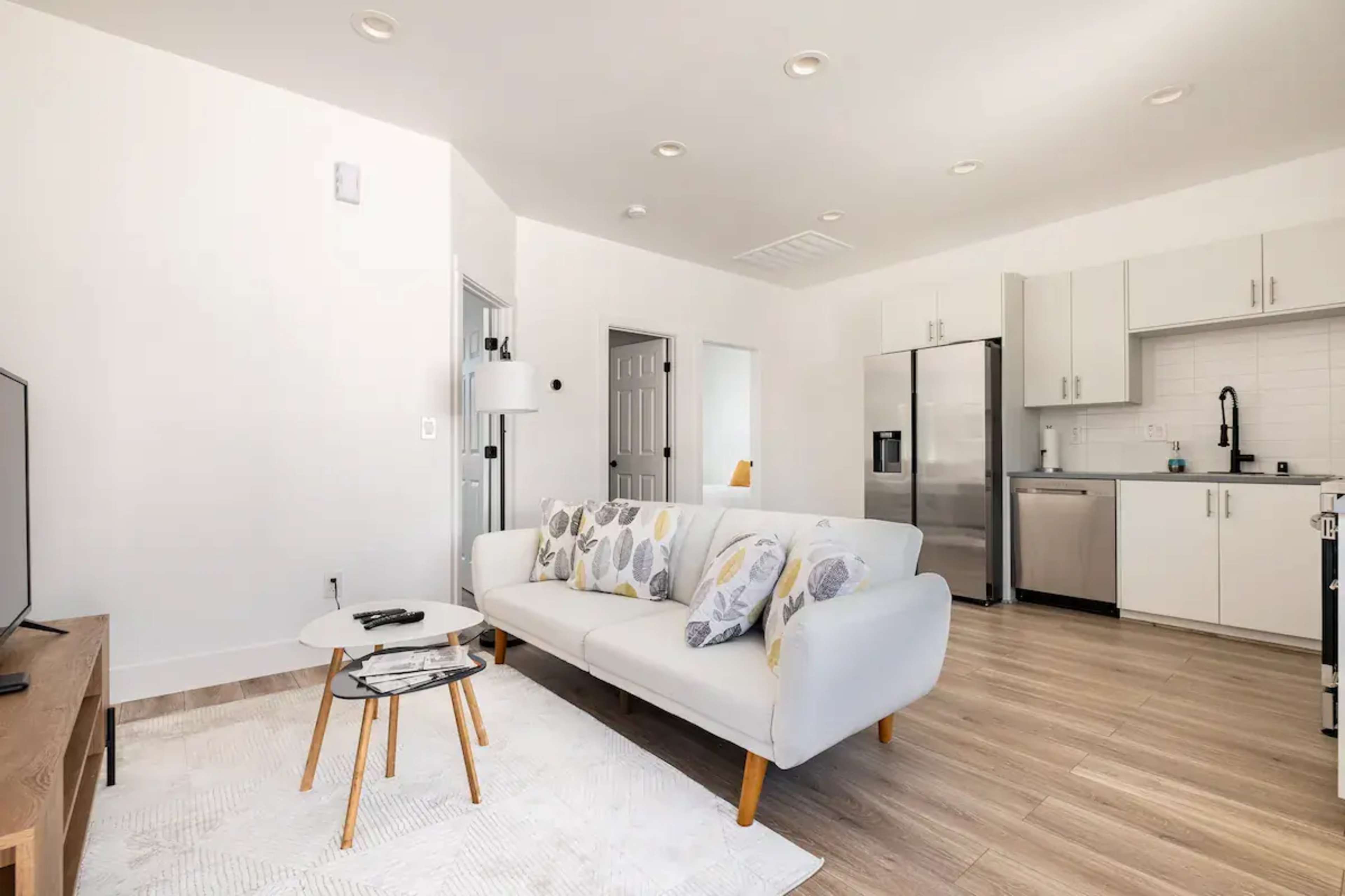 A modern living room features a white sofa with decorative cushions, a small coffee table, and an open kitchen area with stainless steel appliances.