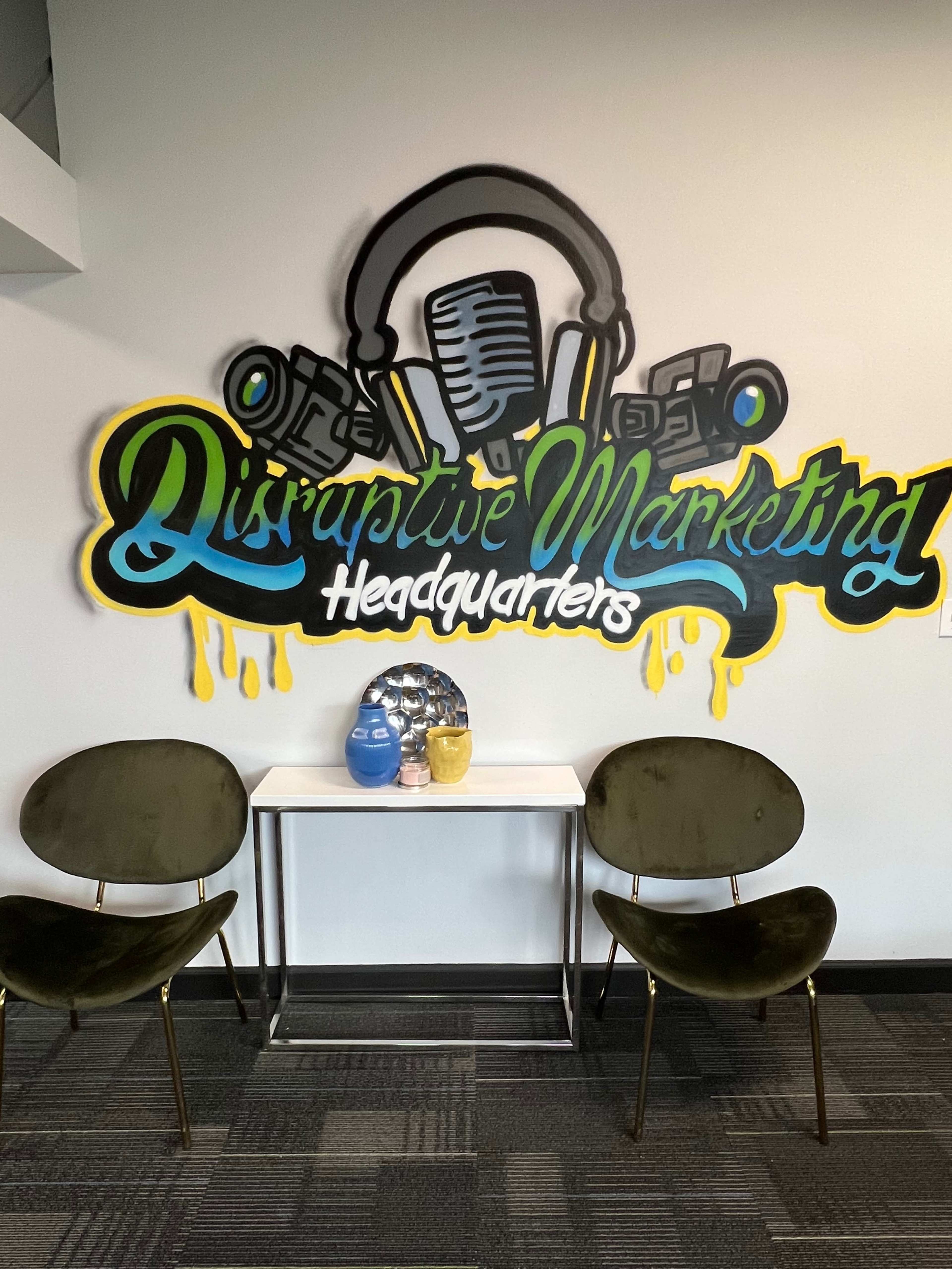 A wall with a colorful mural reading "Disruptive Marketing Headquarters" above a small table and two green chairs.