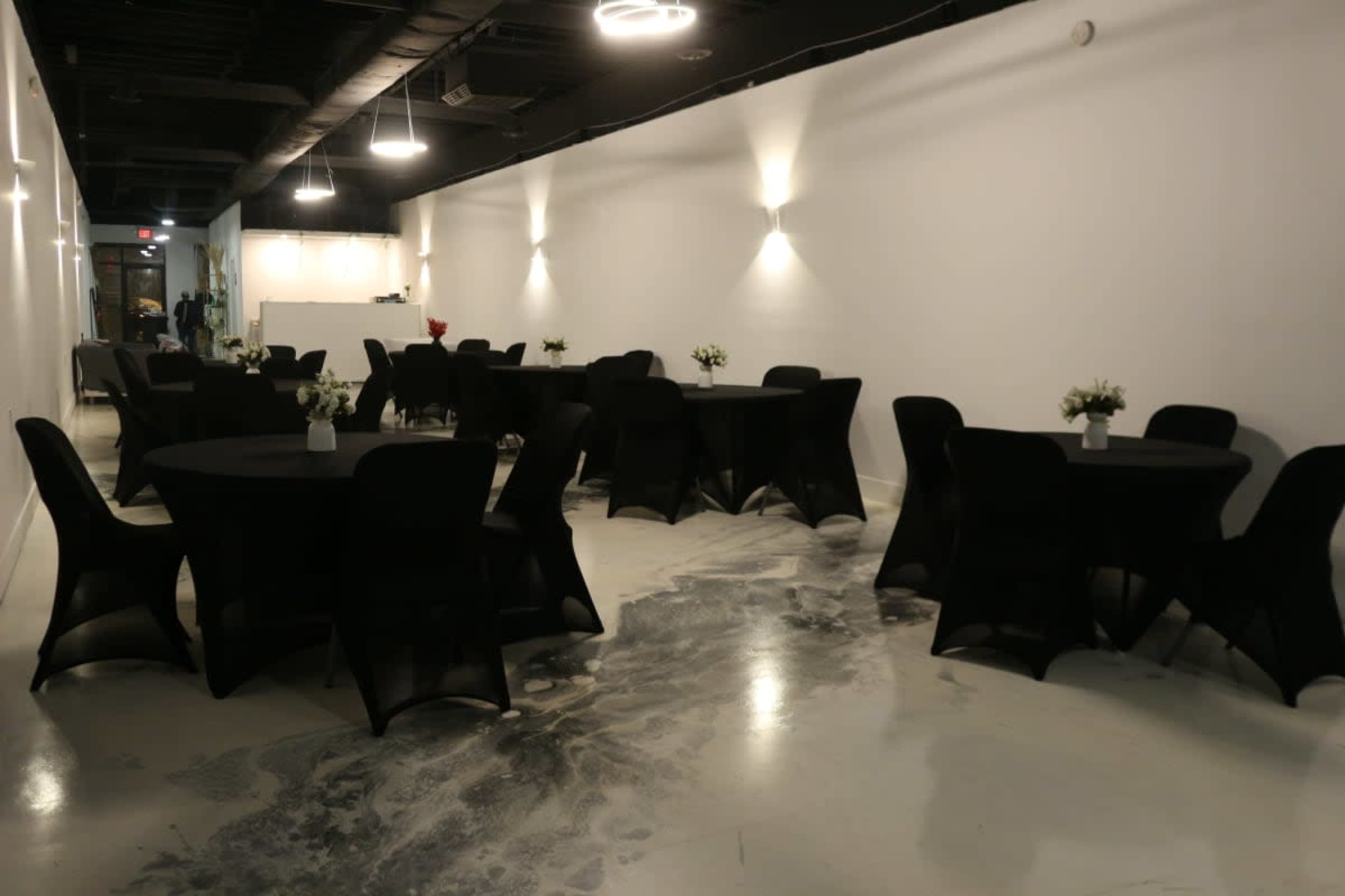 A modern event space with round tables covered in black tablecloths and small floral centerpieces, against a backdrop of white walls and subtle lighting.