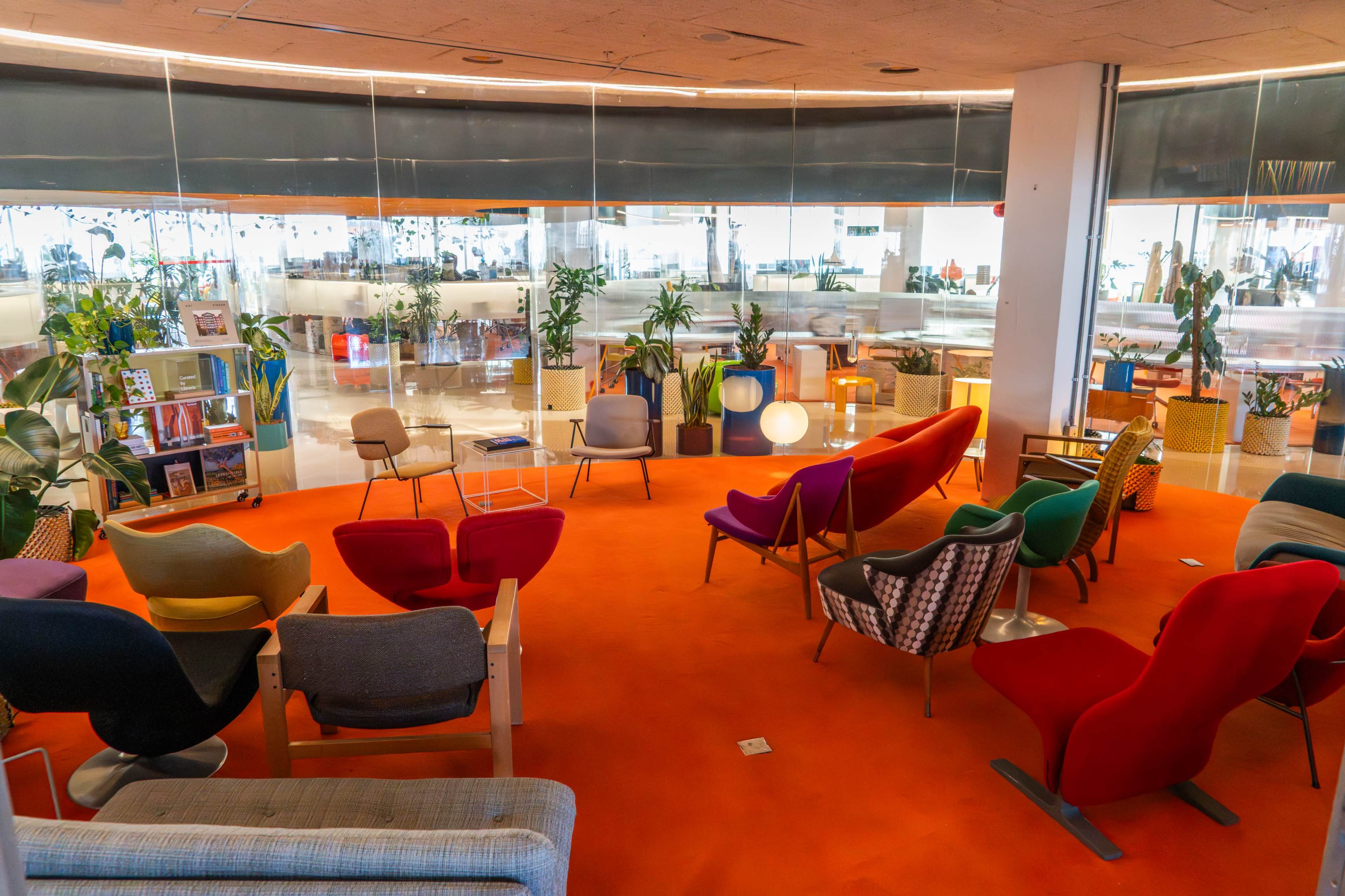 The image shows a modern seating area with various colorful chairs arranged around a vibrant orange carpet in a spacious room filled with plants and natural light.