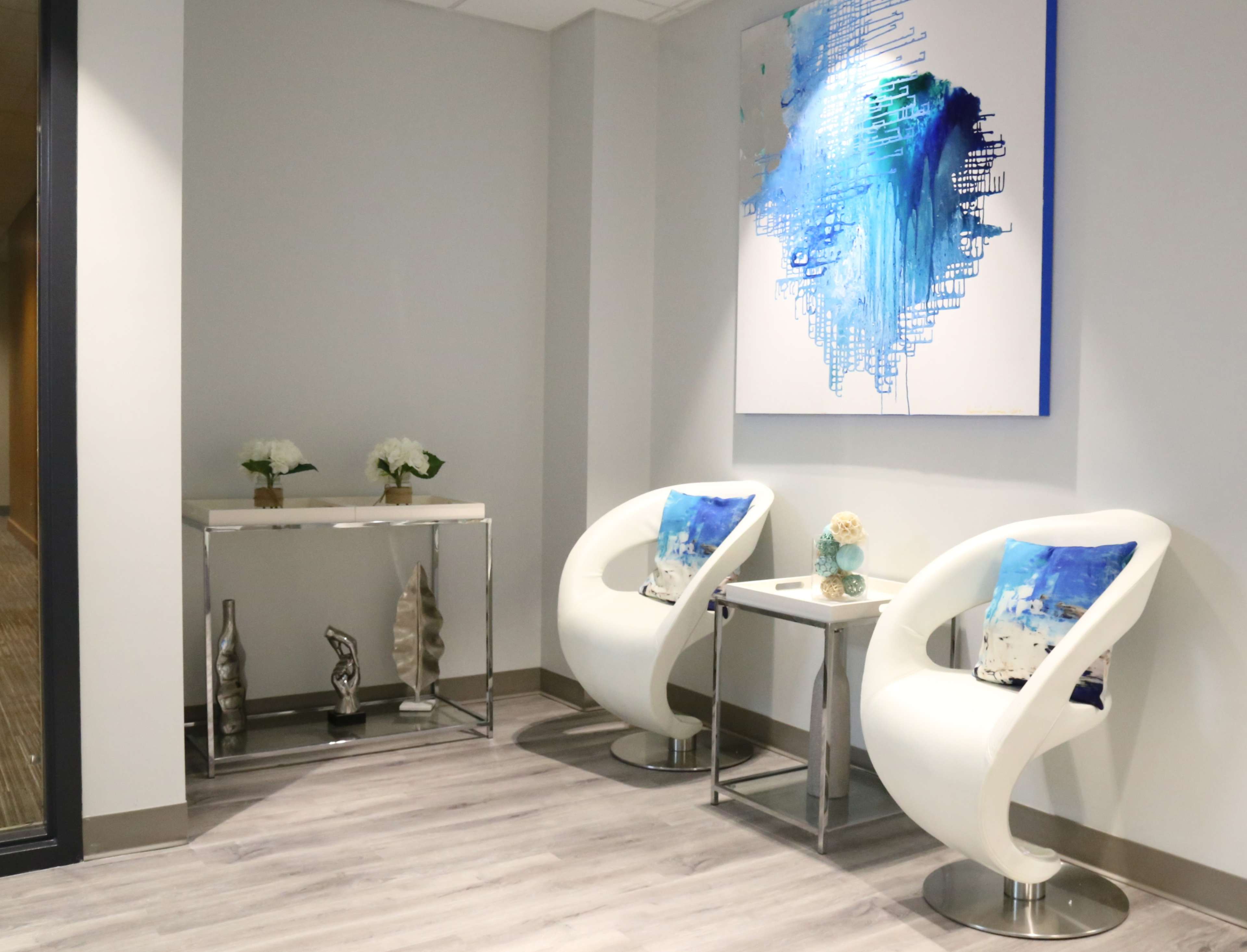 A modern corner of an office lobby featuring two white chairs, a small table, a decorative console table, and a large abstract painting on the wall.