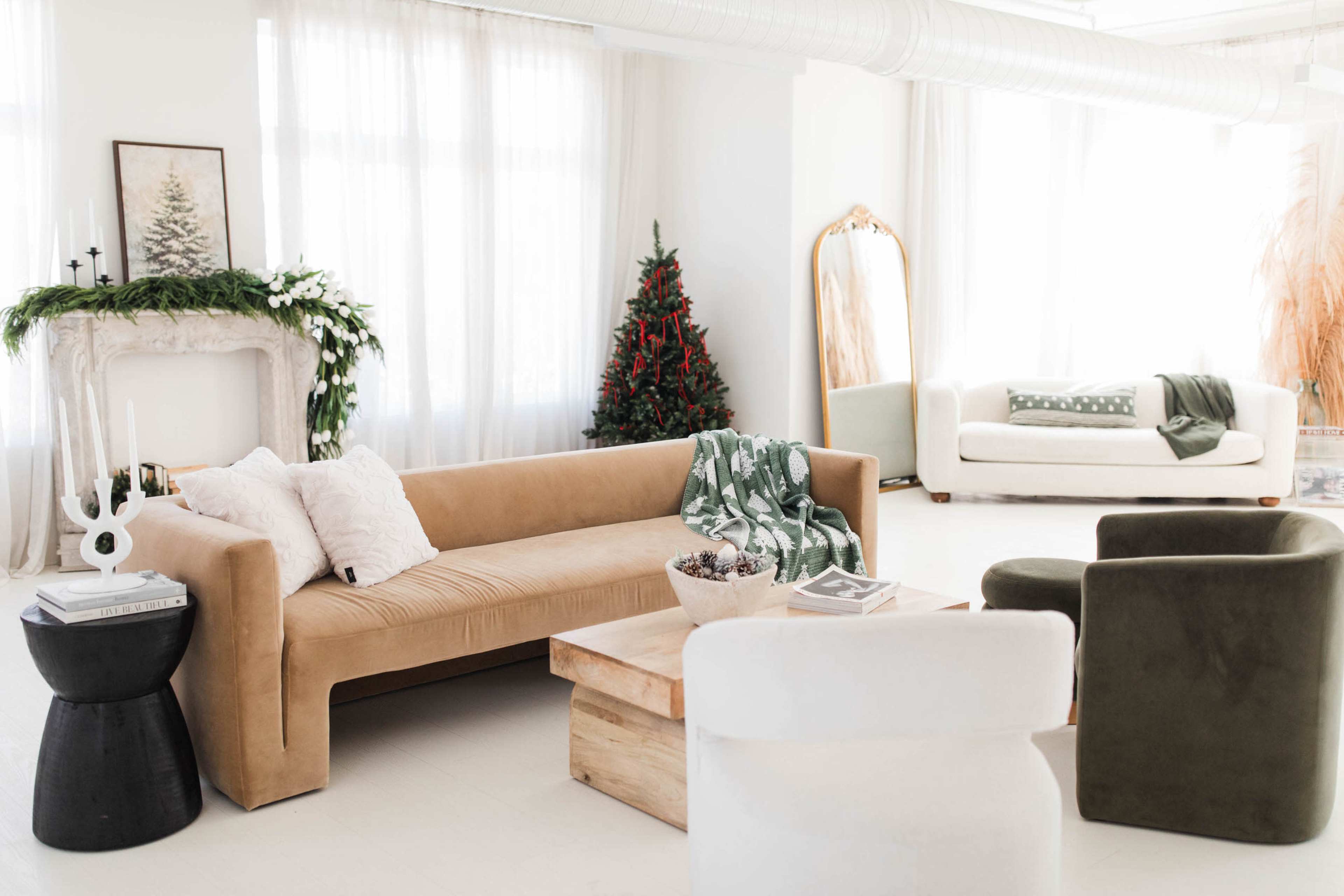 The image shows a bright and airy living room with a beige sofa, a green tree in the corner, and a wooden coffee table surrounded by various chairs.