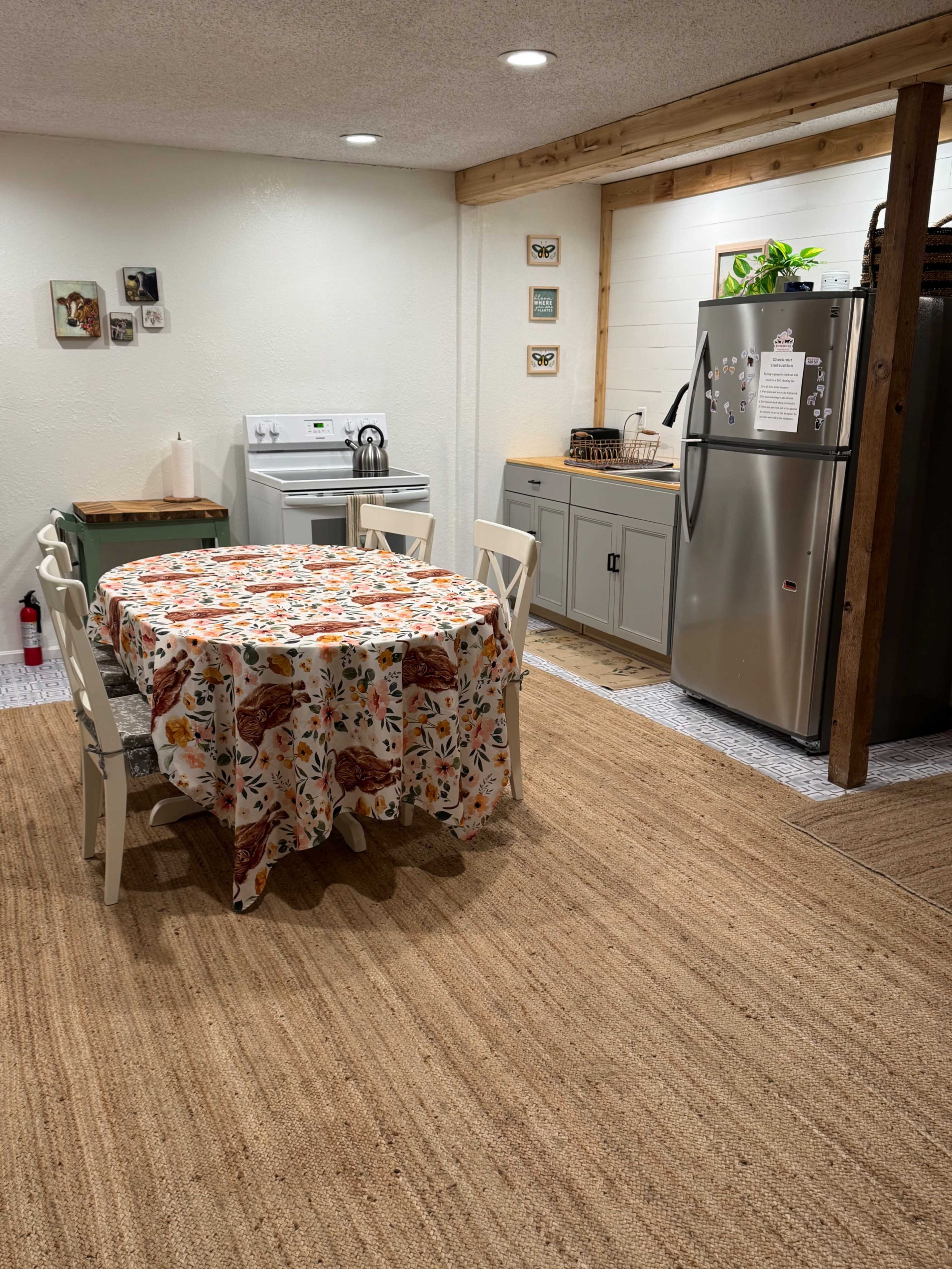 The image shows a kitchen with a round table covered by a patterned tablecloth, surrounded by four chairs, and featuring a stove, refrigerator, and cabinets along the walls.