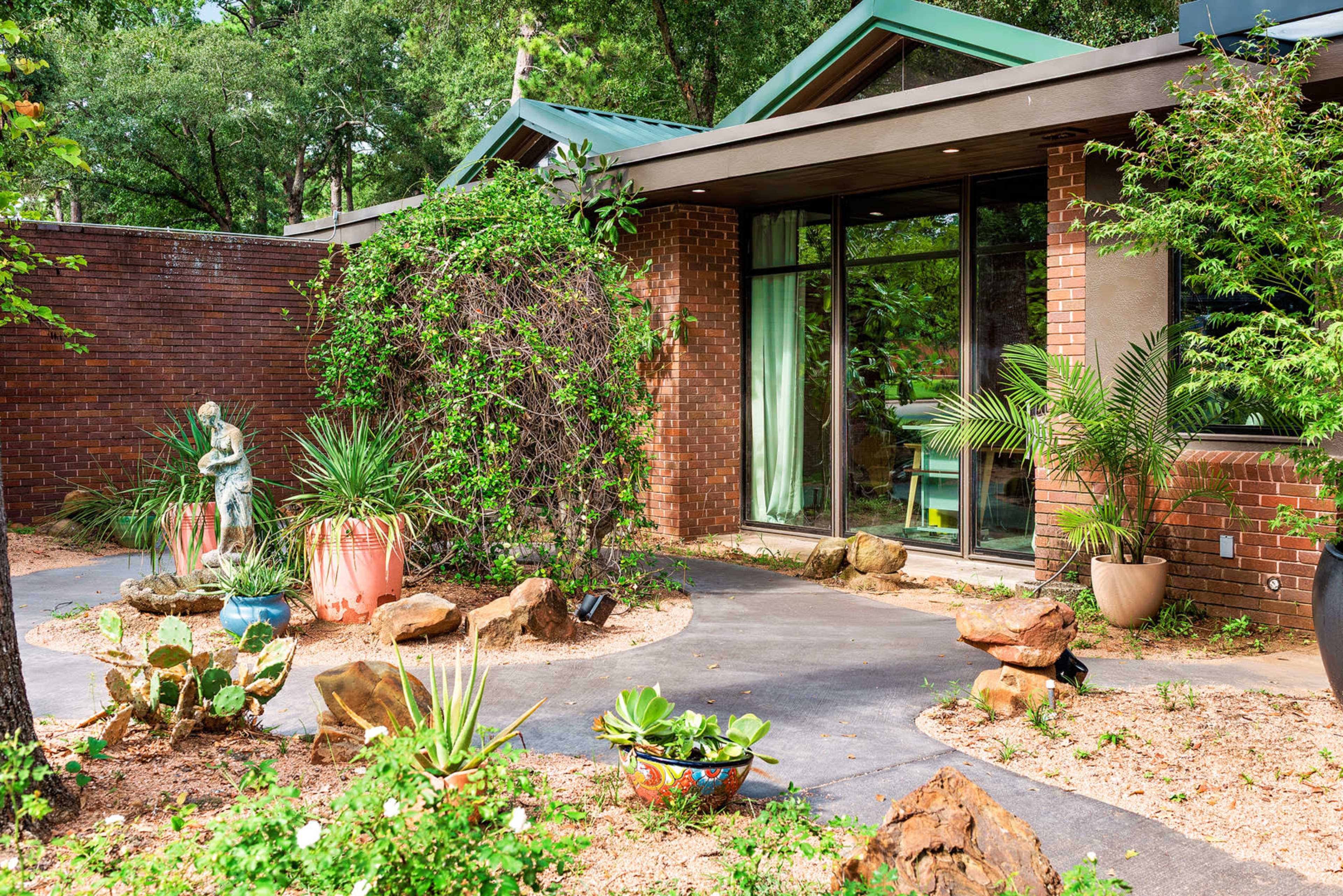 The image shows a modern brick house with a landscaped garden featuring various plants and rock formations, along with a statue in the foreground.