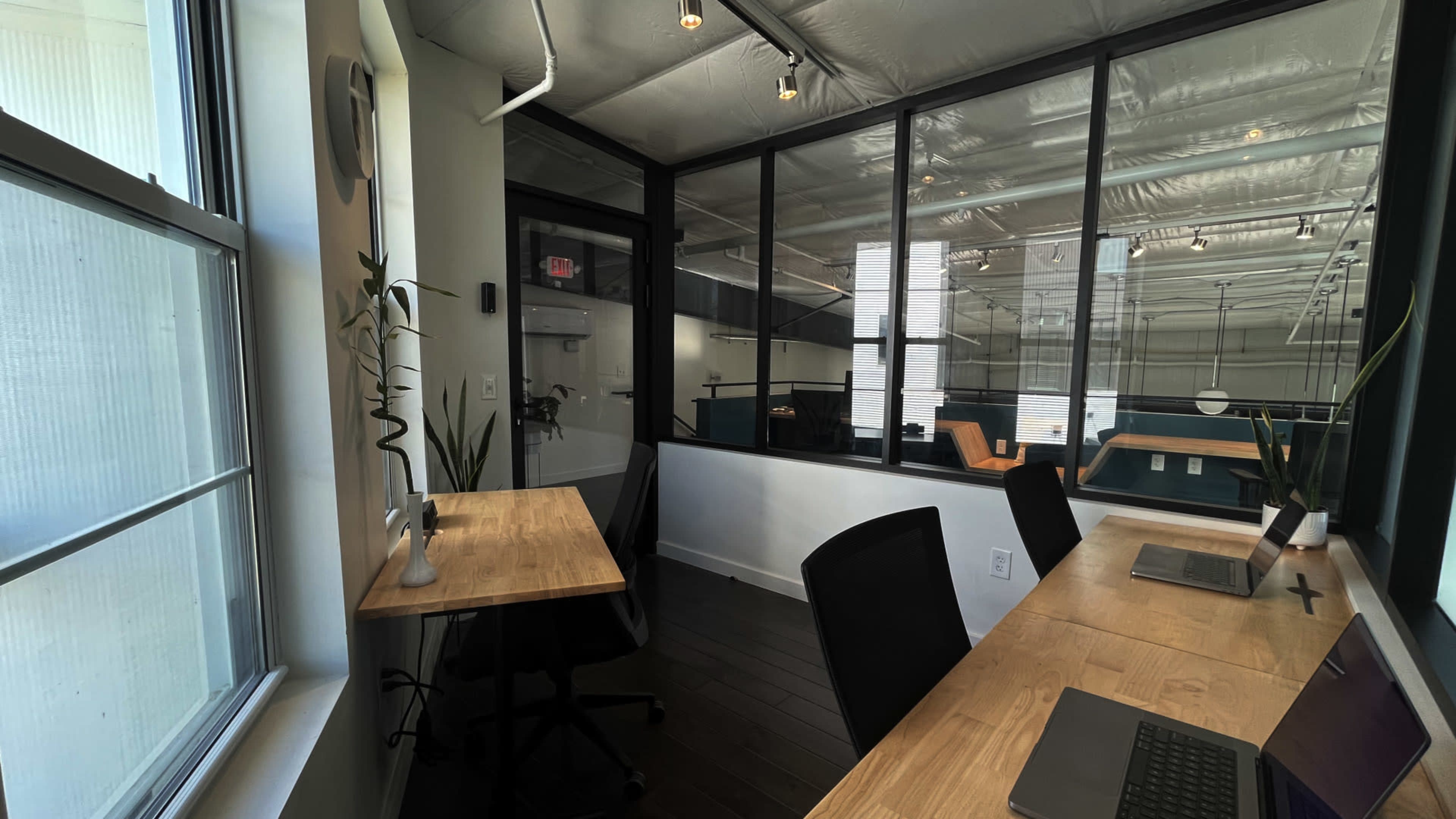 The image shows a modern office space with large windows, a wooden desk, and ergonomic chairs, featuring an open area visible through a glass partition.