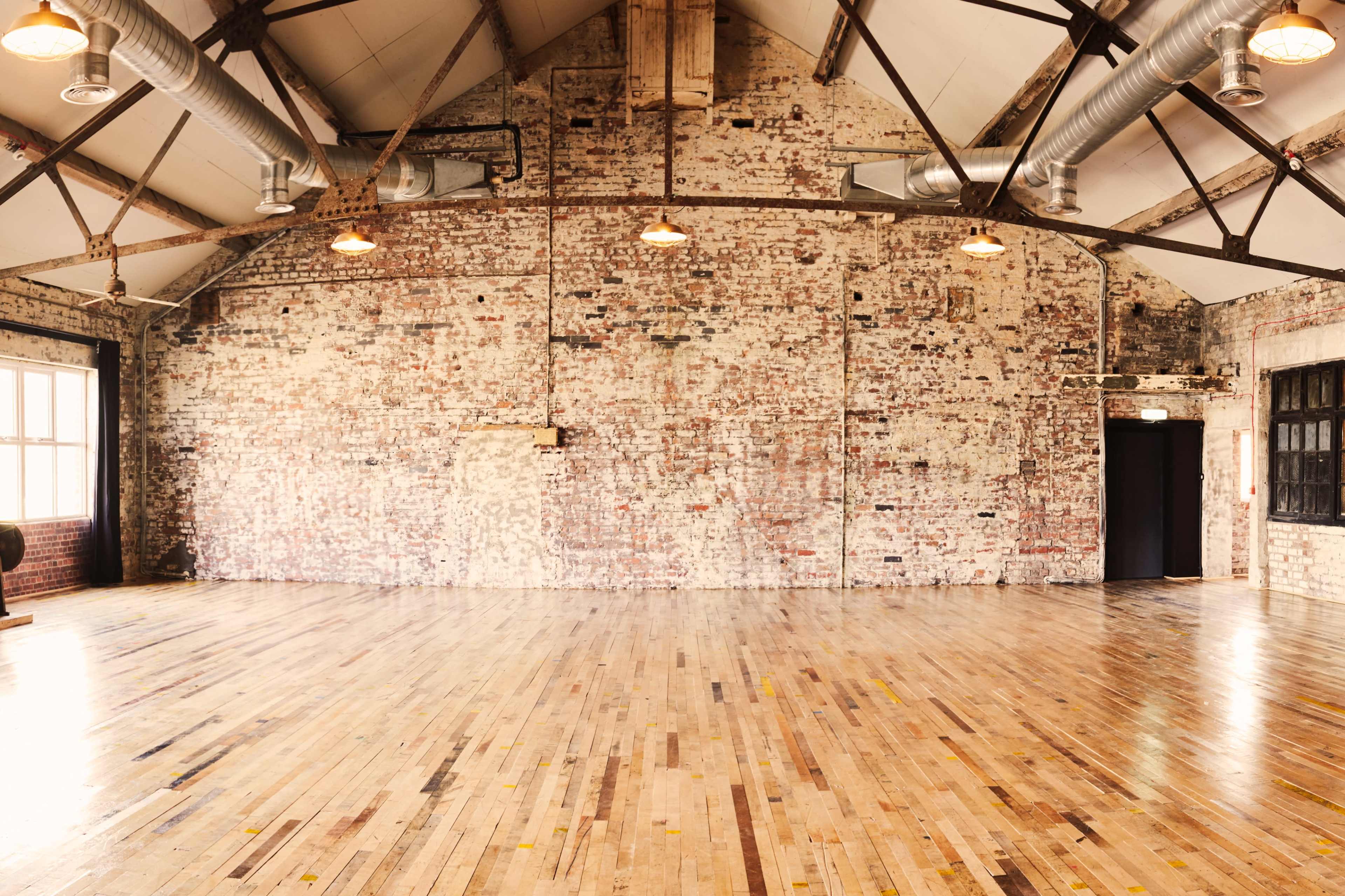 A spacious, industrial-style room with exposed brick walls and polished wooden floors, illuminated by pendant lights.