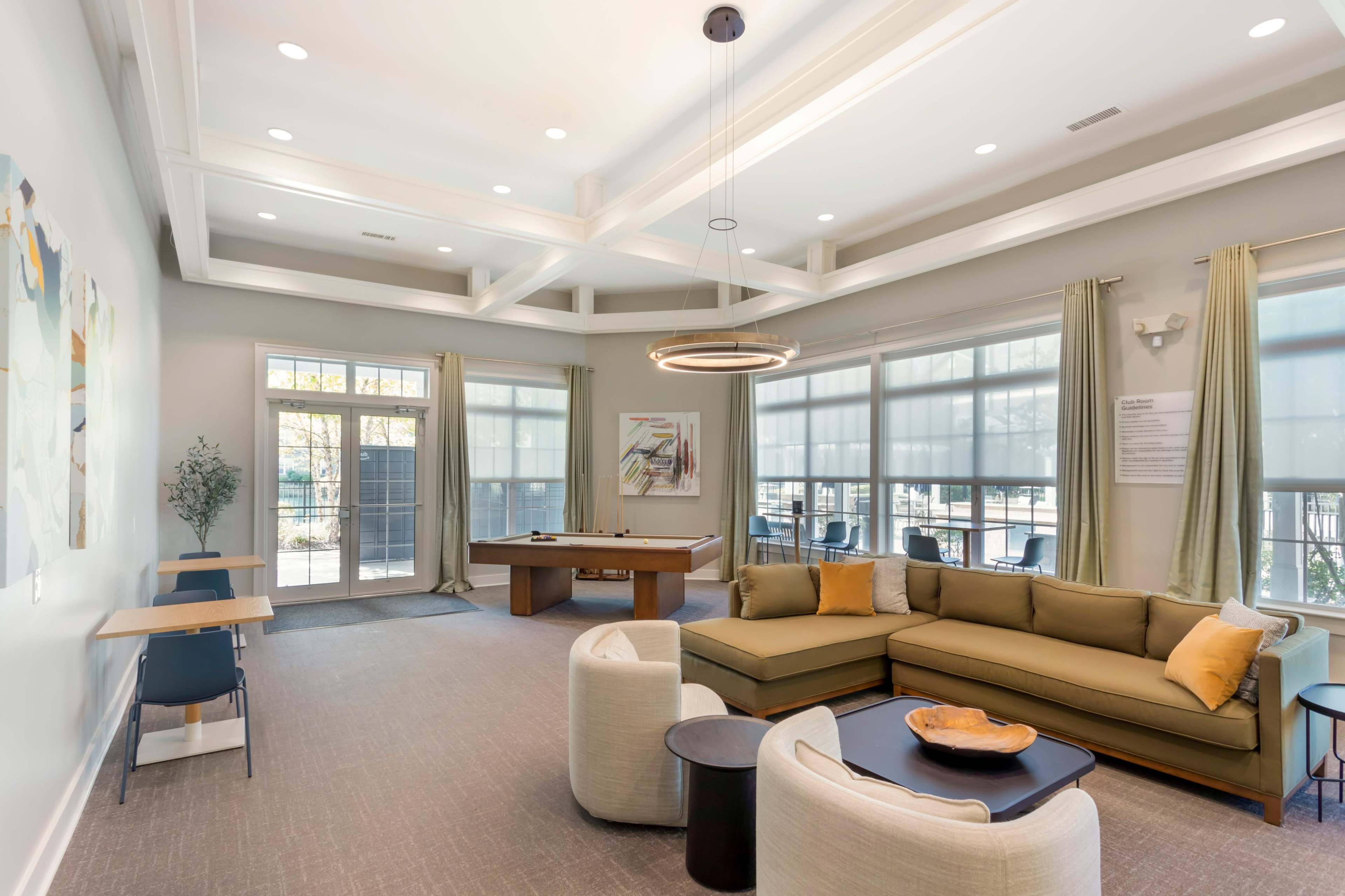 The image shows a spacious community lounge with a sectional sofa, a pool table, and large windows allowing natural light.