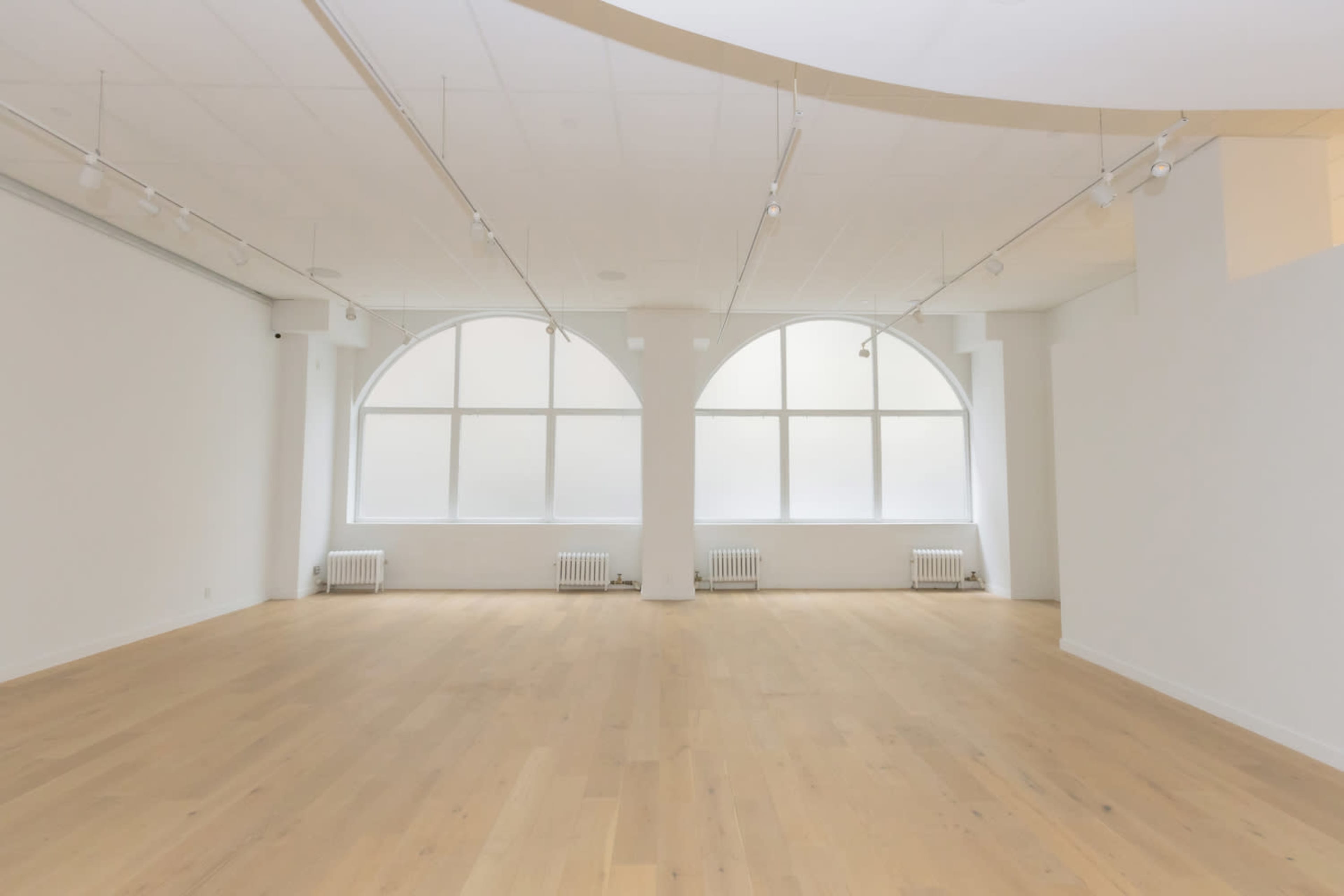 The image shows a bright, empty room with large arch-shaped windows and wooden flooring.