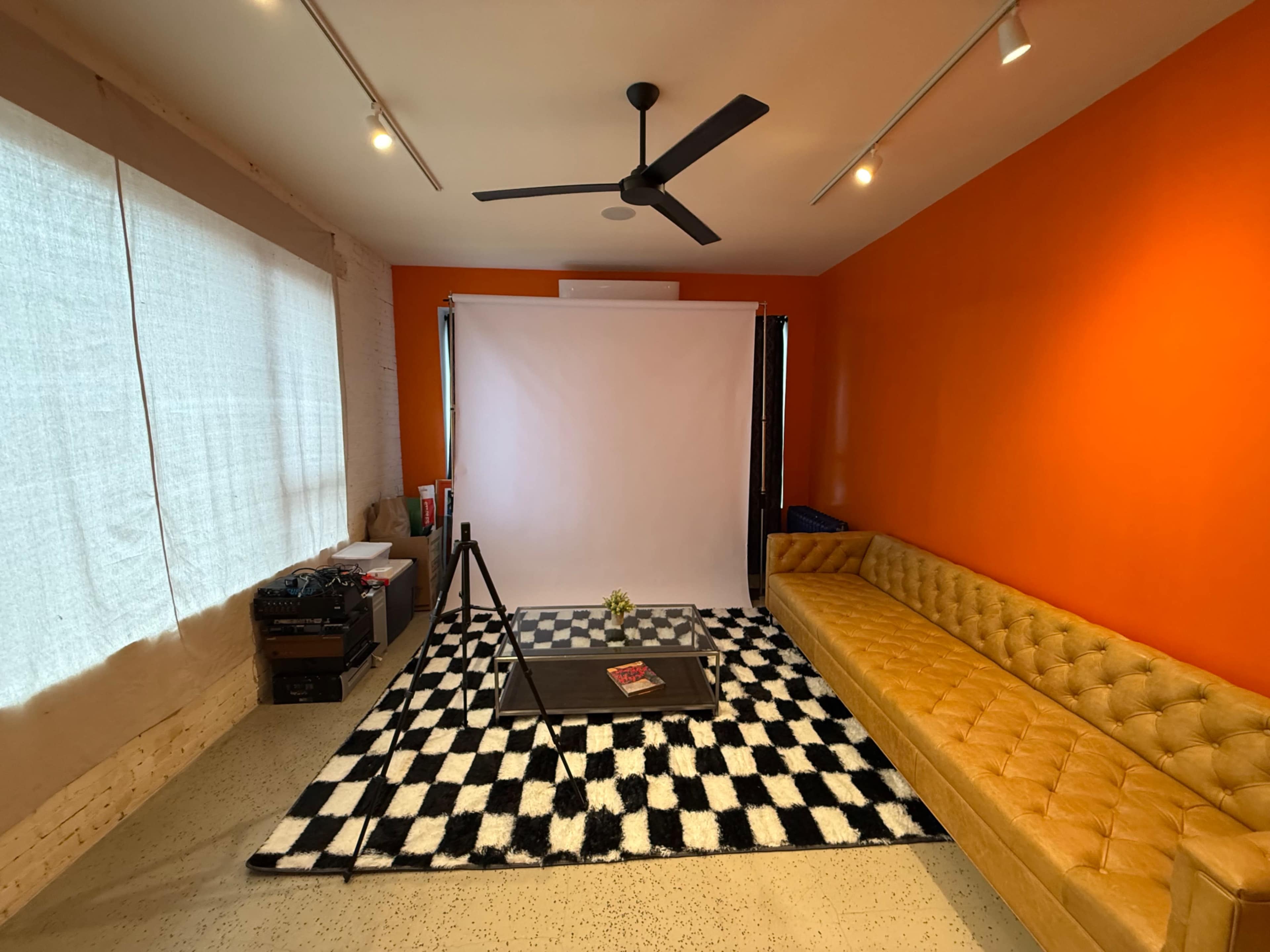 The image shows a modern interior space with an orange accent wall, a yellow tufted sofa, a black and white checkered rug, and a backdrop on a stand set up for photography.