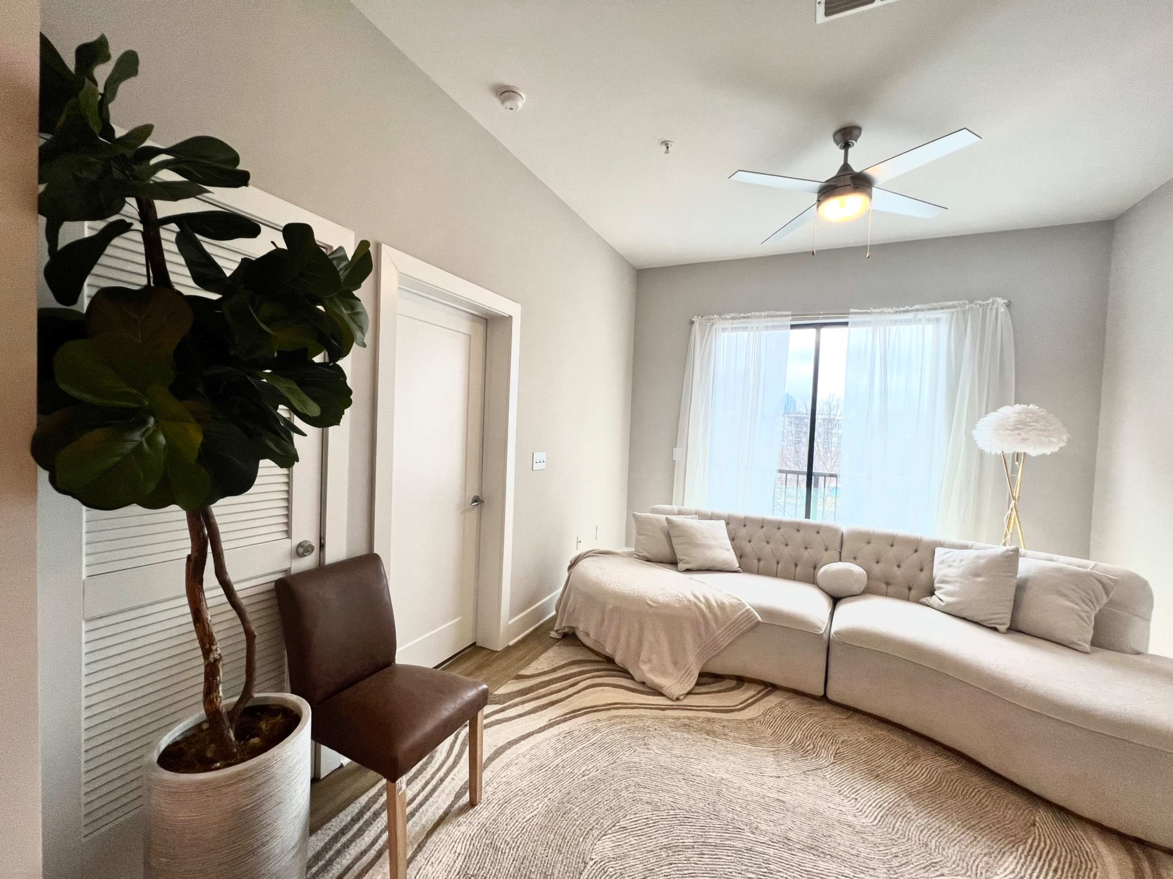 A modern living room featuring a light gray curved sofa, a potted plant, a brown chair, and a spacious window with sheer curtains.