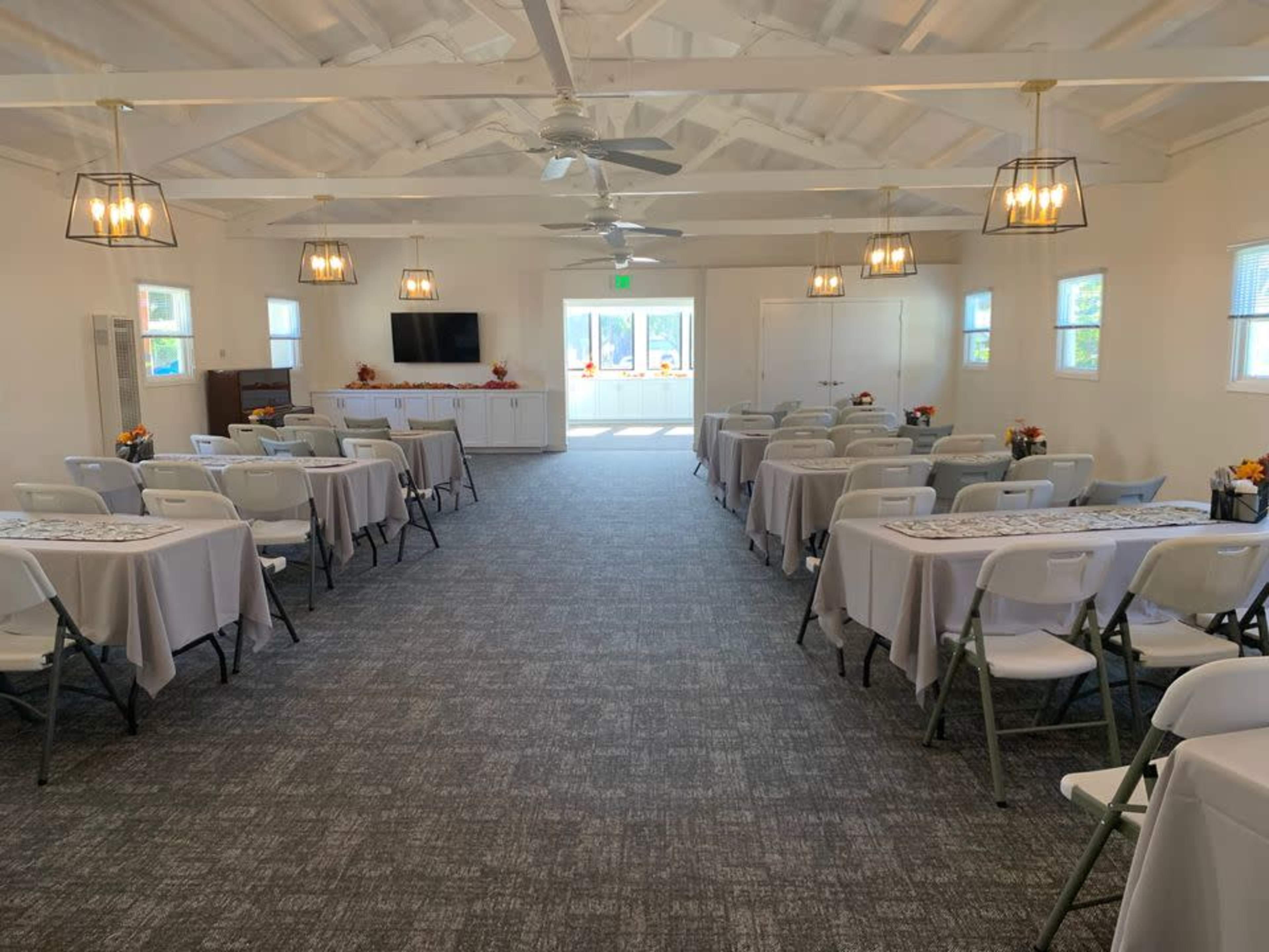 A well-lit meeting room with several tables arranged in rows, each covered with a tablecloth, and features large windows and a television at the front.
