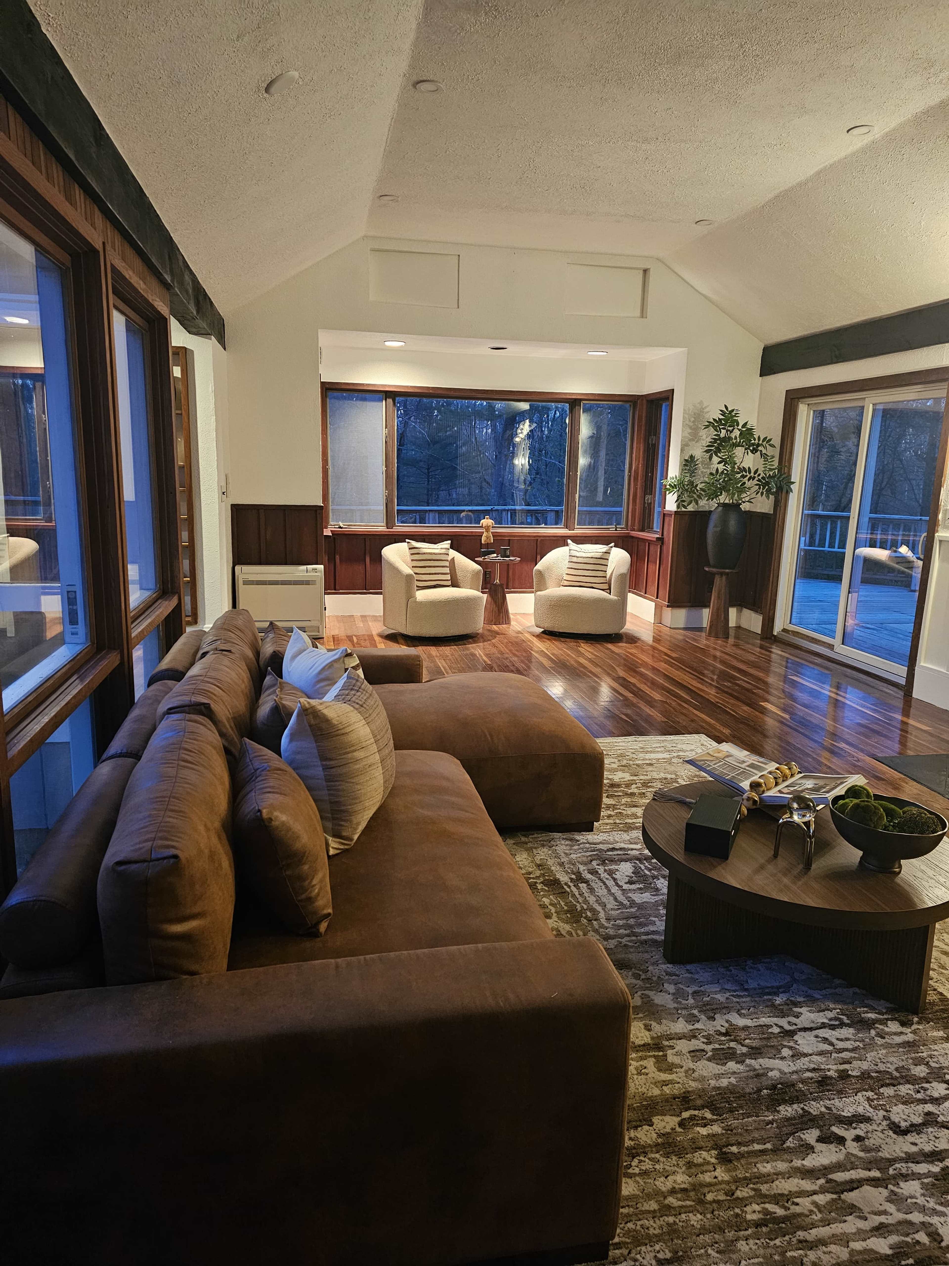 The image shows a modern living room featuring a brown sectional sofa, two armchairs, and large windows overlooking a wooded area.