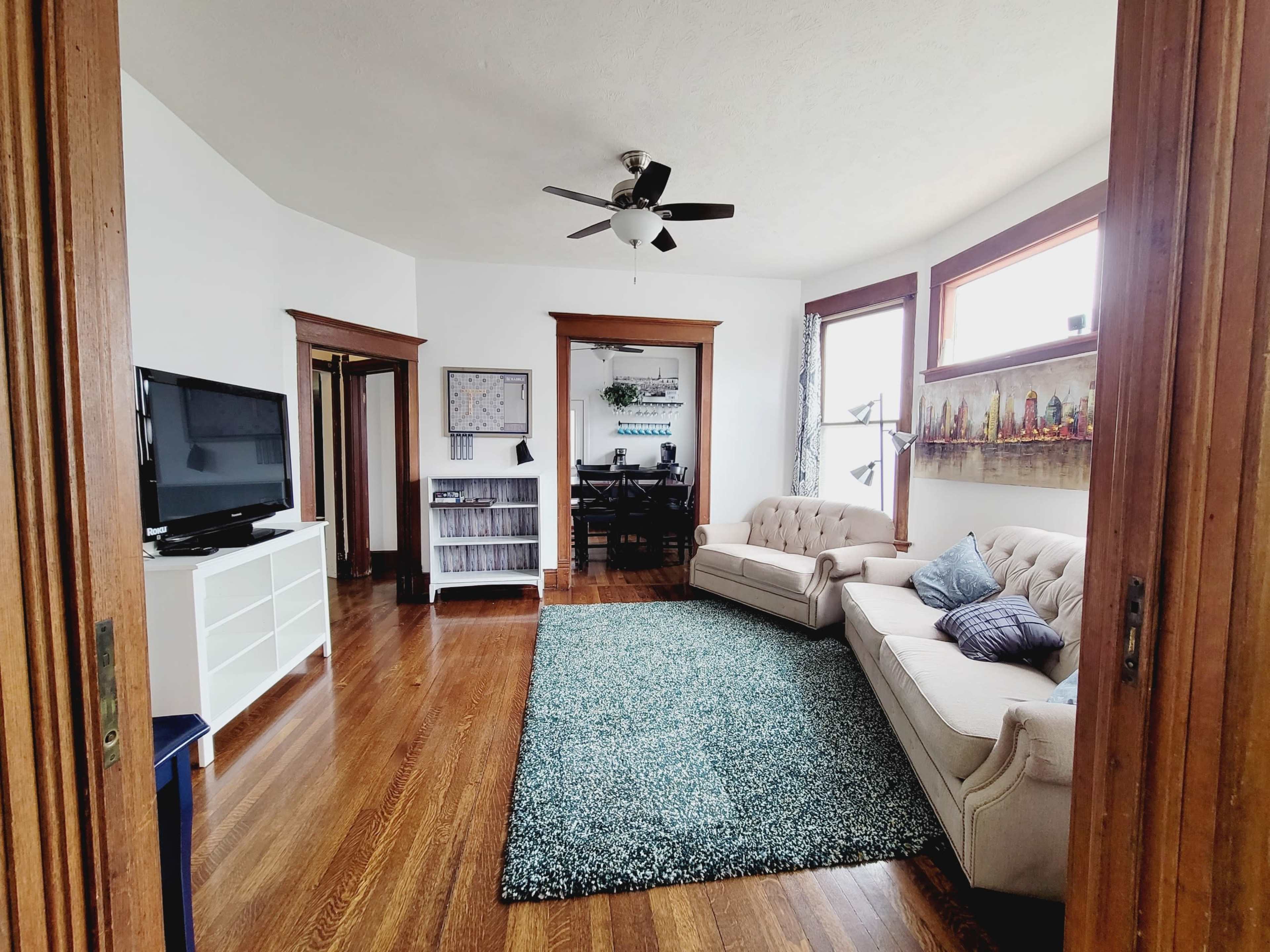 A living room with a beige couch, a television on a white stand, and a dining area visible in the background.