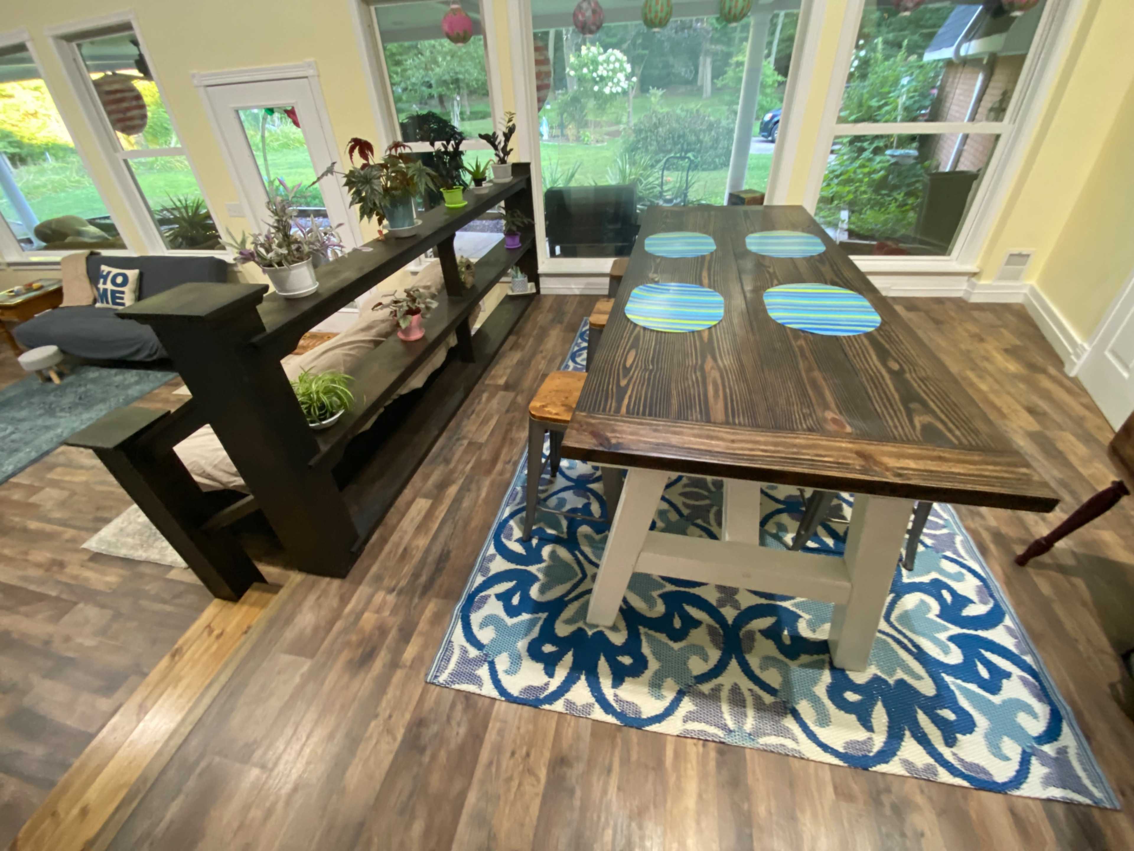 A room with a wooden dining table featuring blue-striped placemats, surrounded by a patterned rug, with a black shelf displaying plants and a seating area in the background.