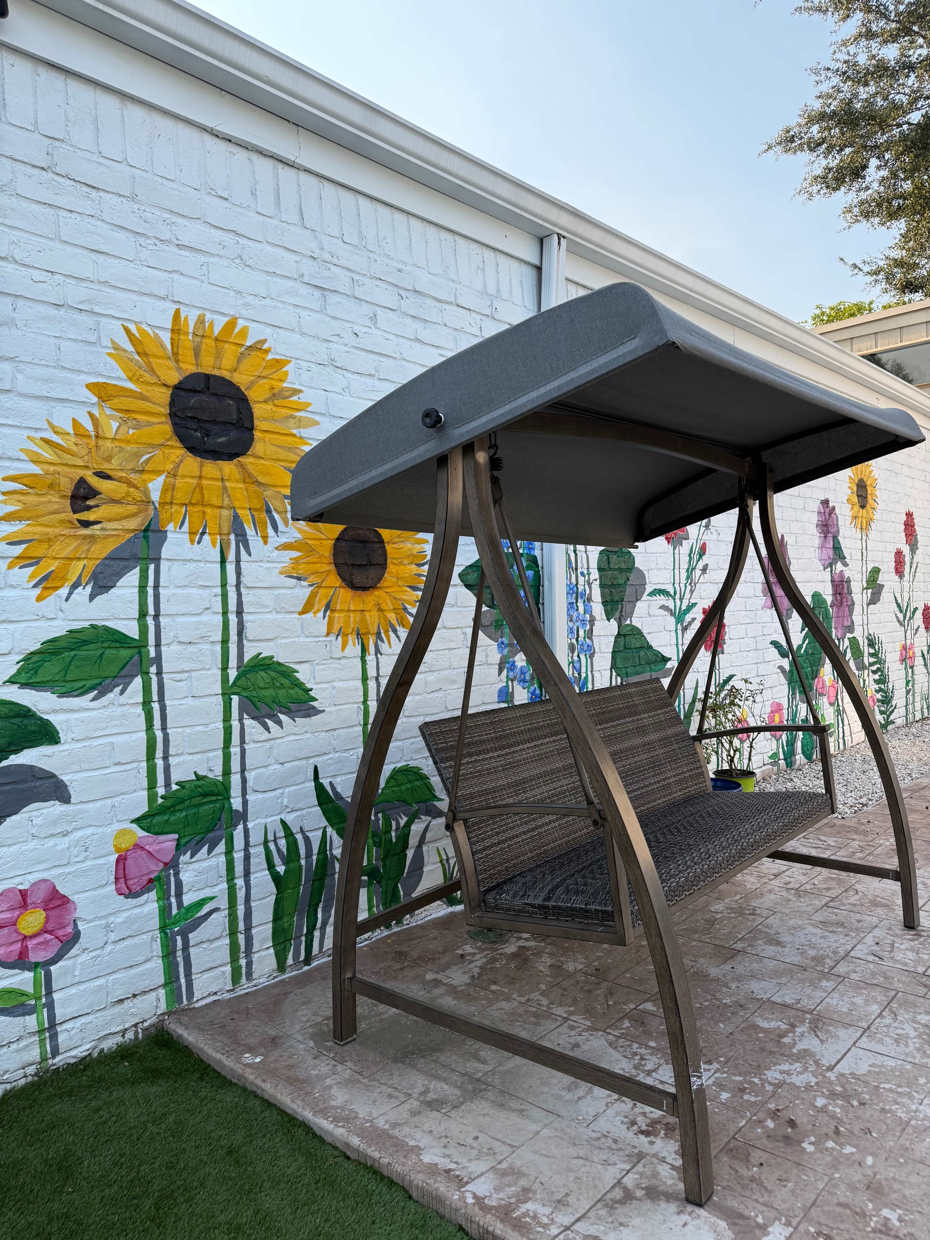 A swing bench is positioned in front of a vibrant mural featuring sunflowers and various flowers on a white brick wall.