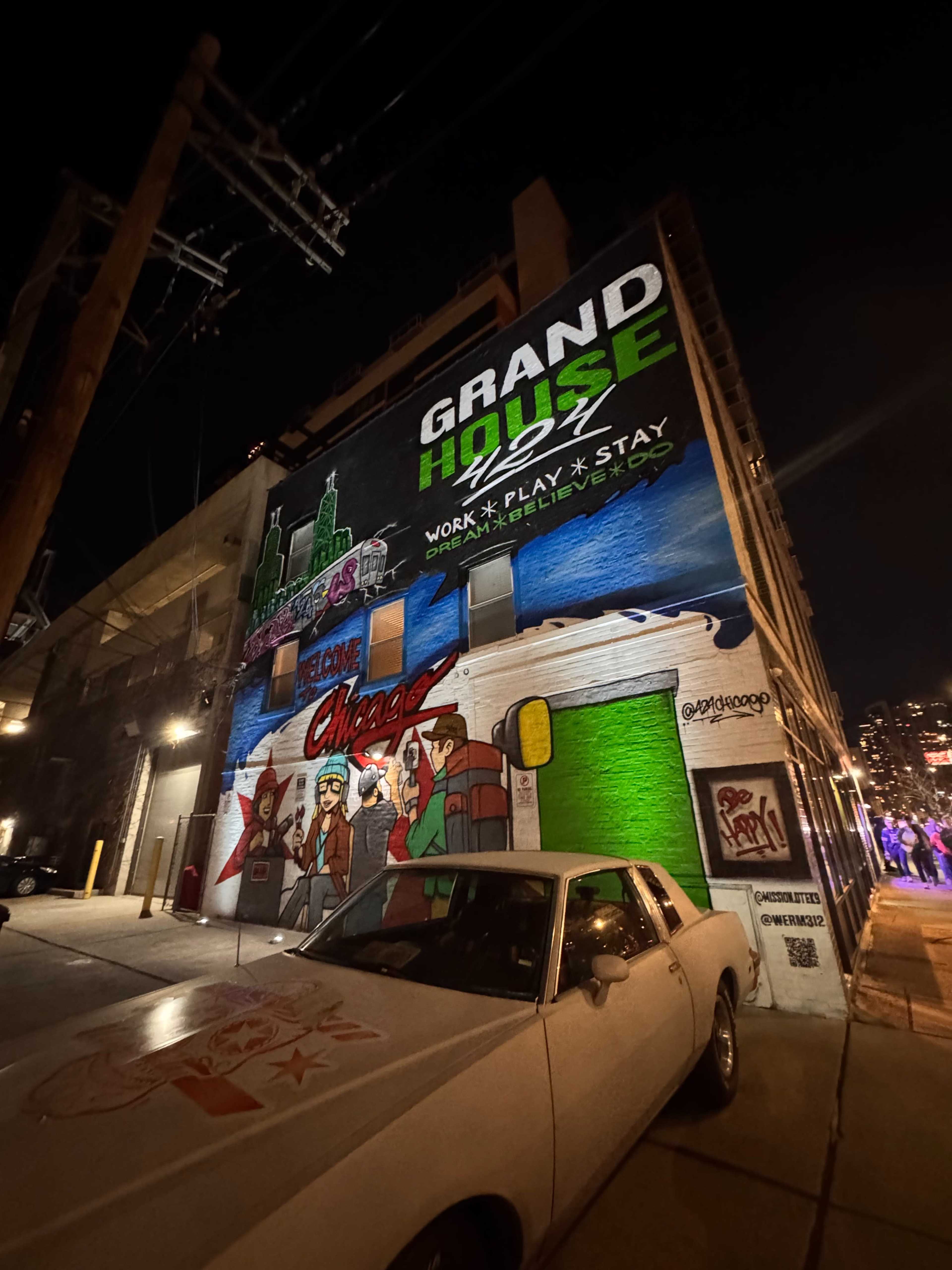 A large mural on the side of a building features the words "GRAND HOUSE" and "WORK • PLAY • STAY," accompanied by images representing Chicago nightlife, alongside a parked vintage car.