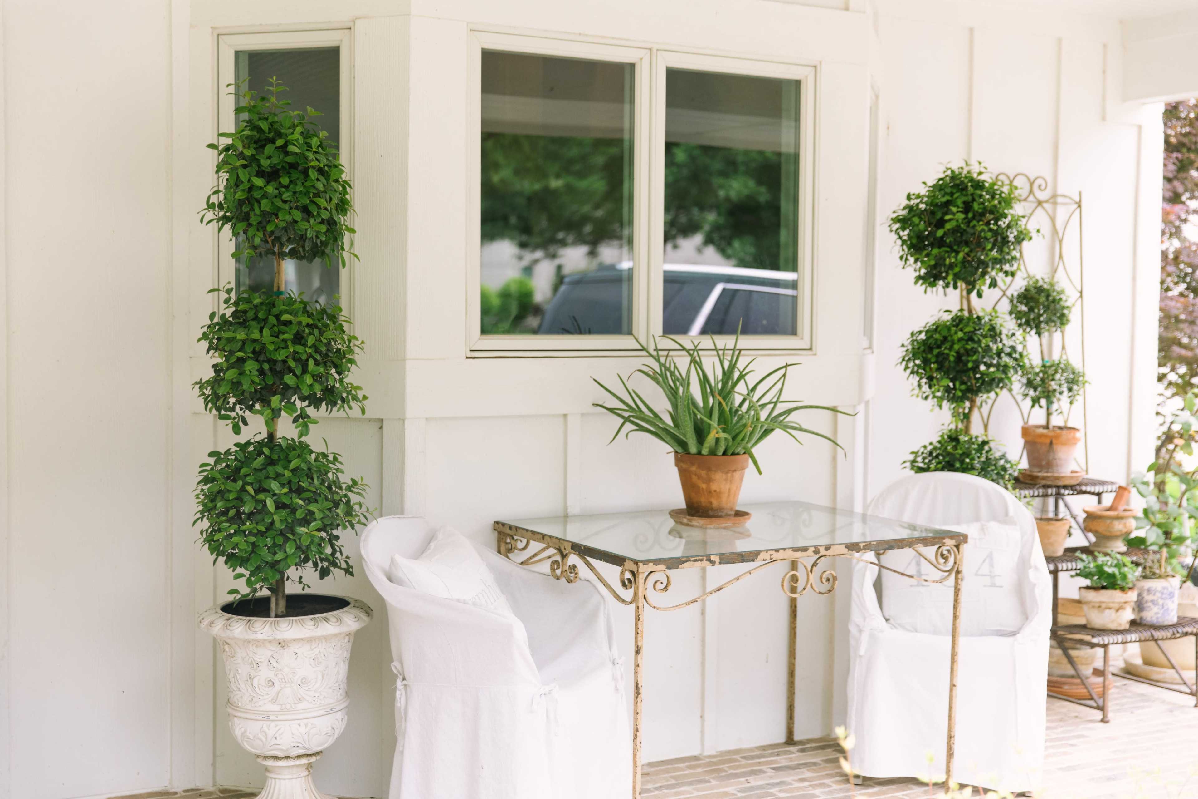 The image shows a cozy porch area featuring a glass table, two white chairs, and potted plants arranged neatly against a wall.