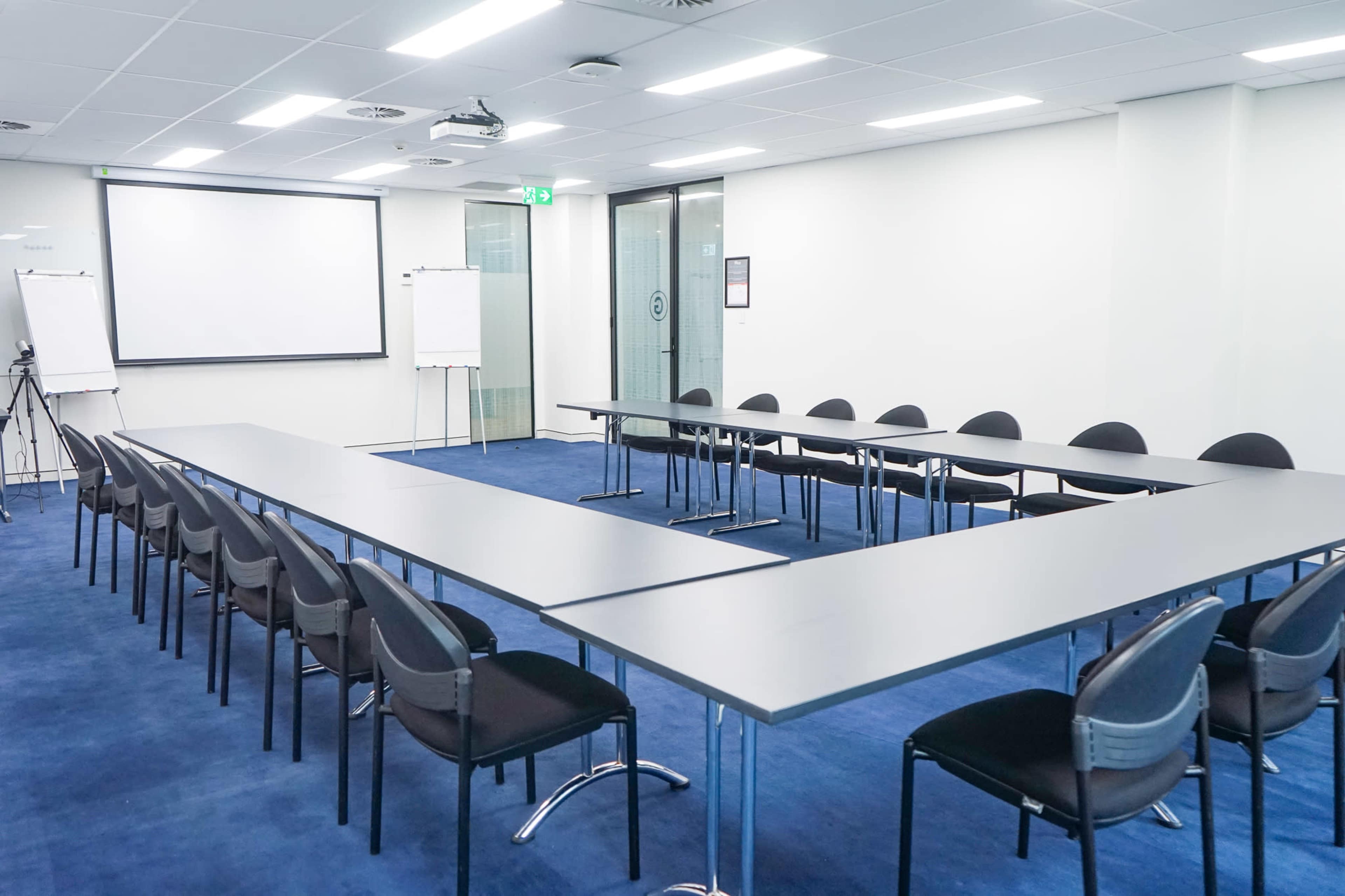 A spacious conference room features a long arrangement of tables and black chairs, with a projector screen and whiteboards at one end.