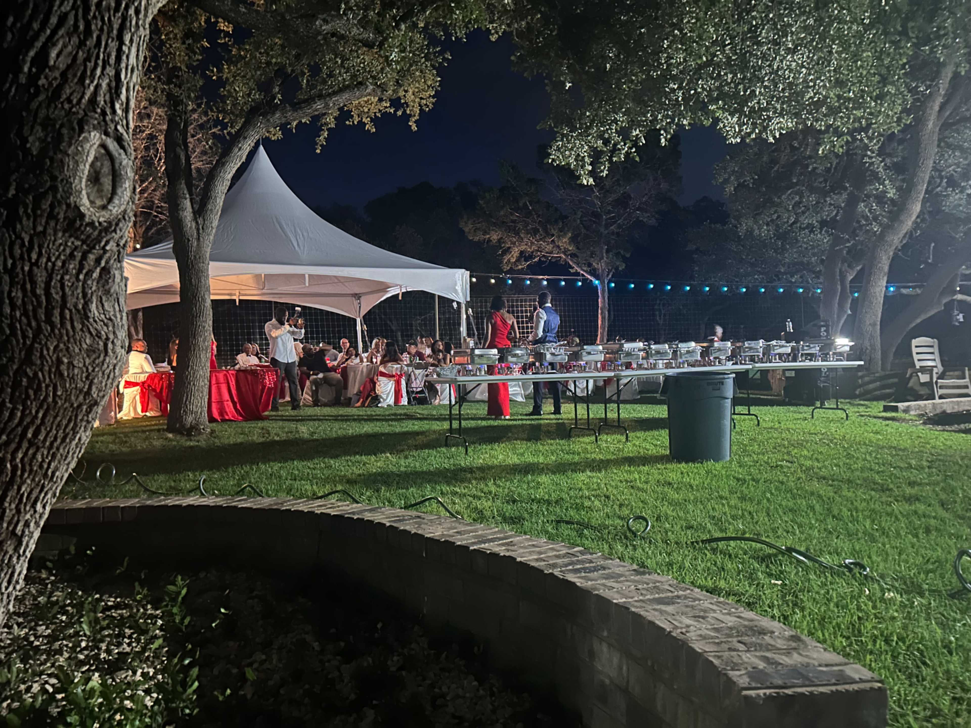 A gathering is taking place under a large white tent in a backyard, with tables set up and people mingling in evening attire.