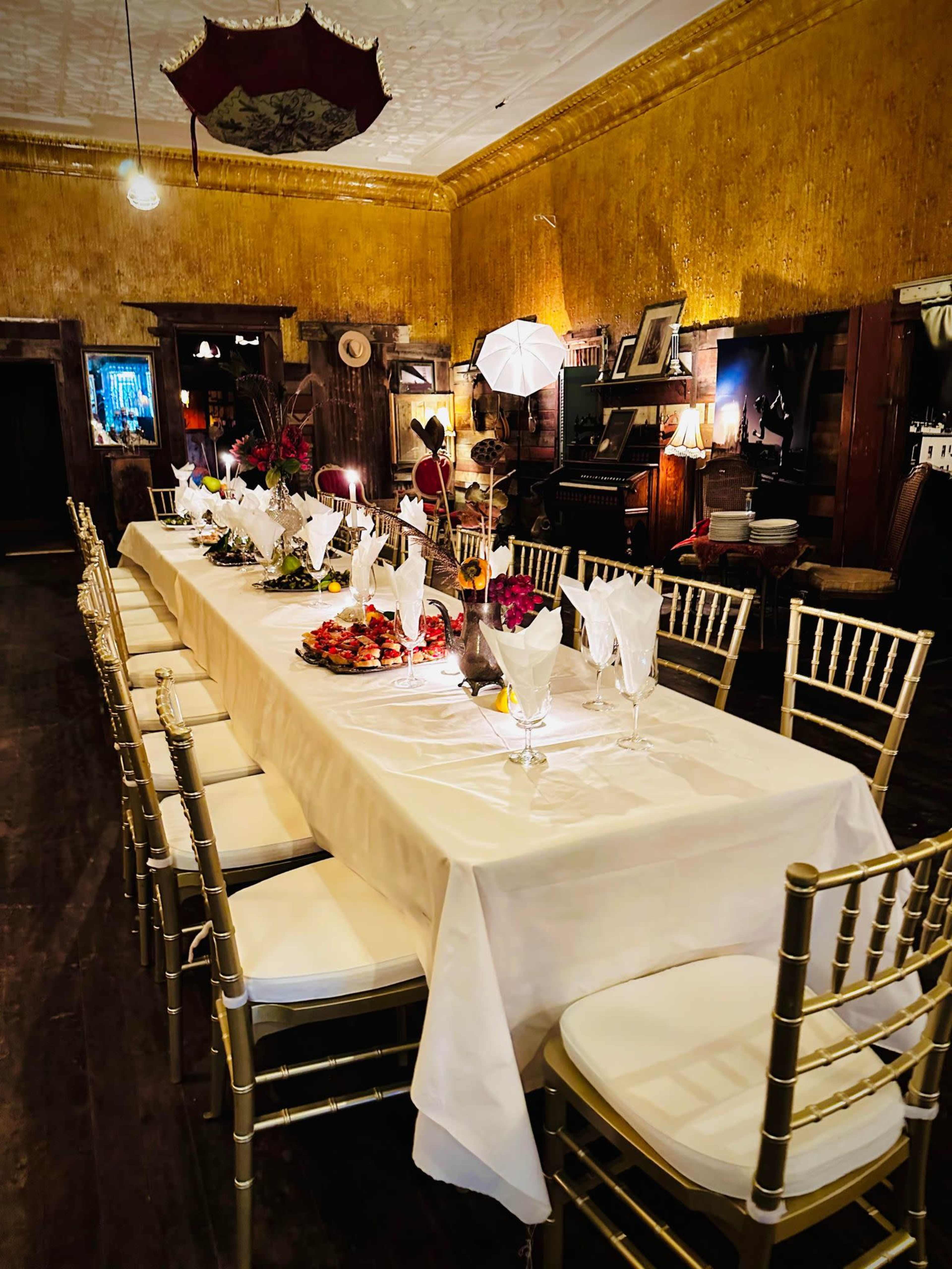 A long, elegantly set dining table adorned with floral arrangements and white tableware is positioned in a vintage room with gold-patterned walls and antique decorations.