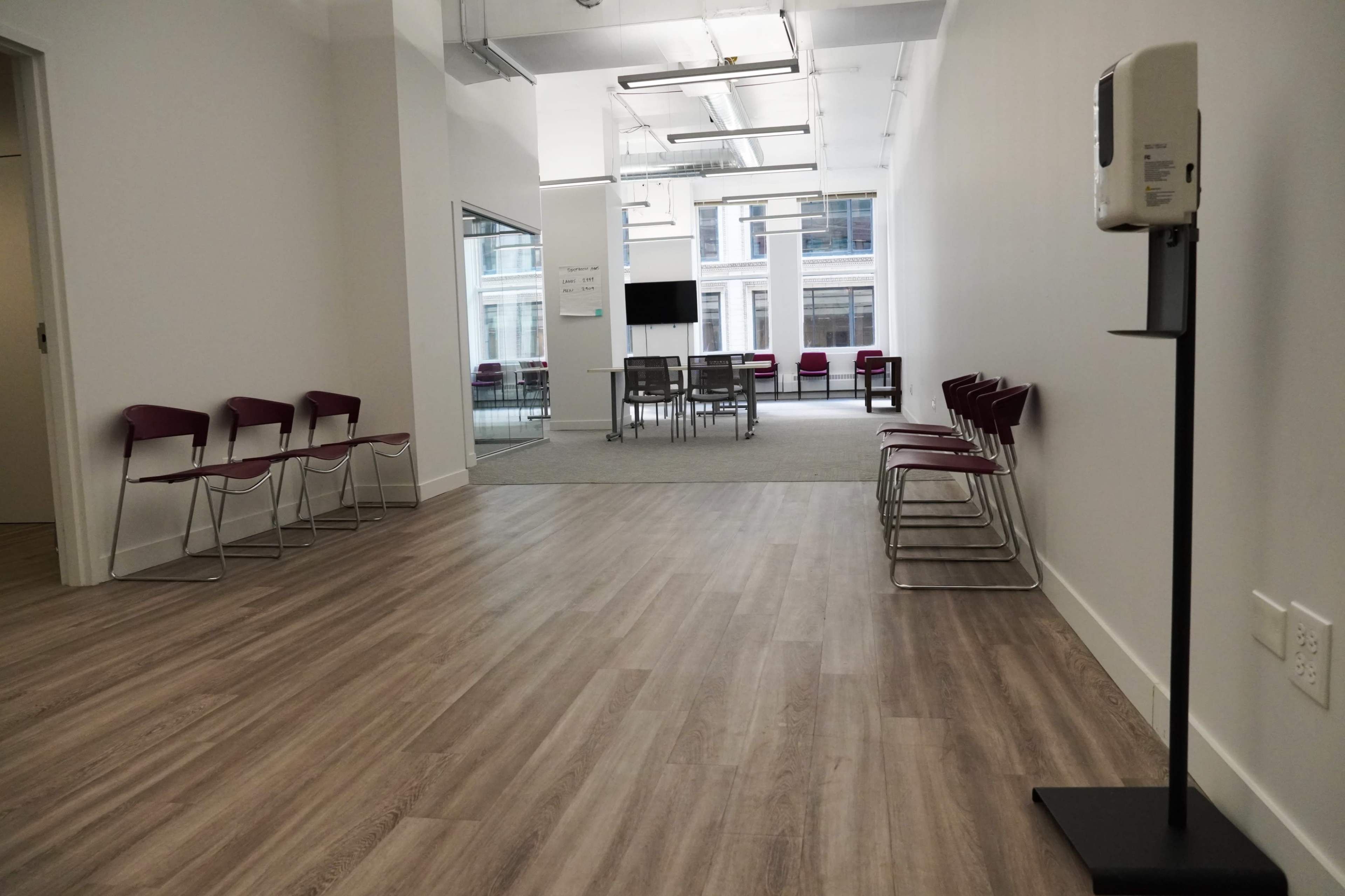 A minimalist waiting area features a row of metal chairs on one side and a glass-walled meeting space in the background.