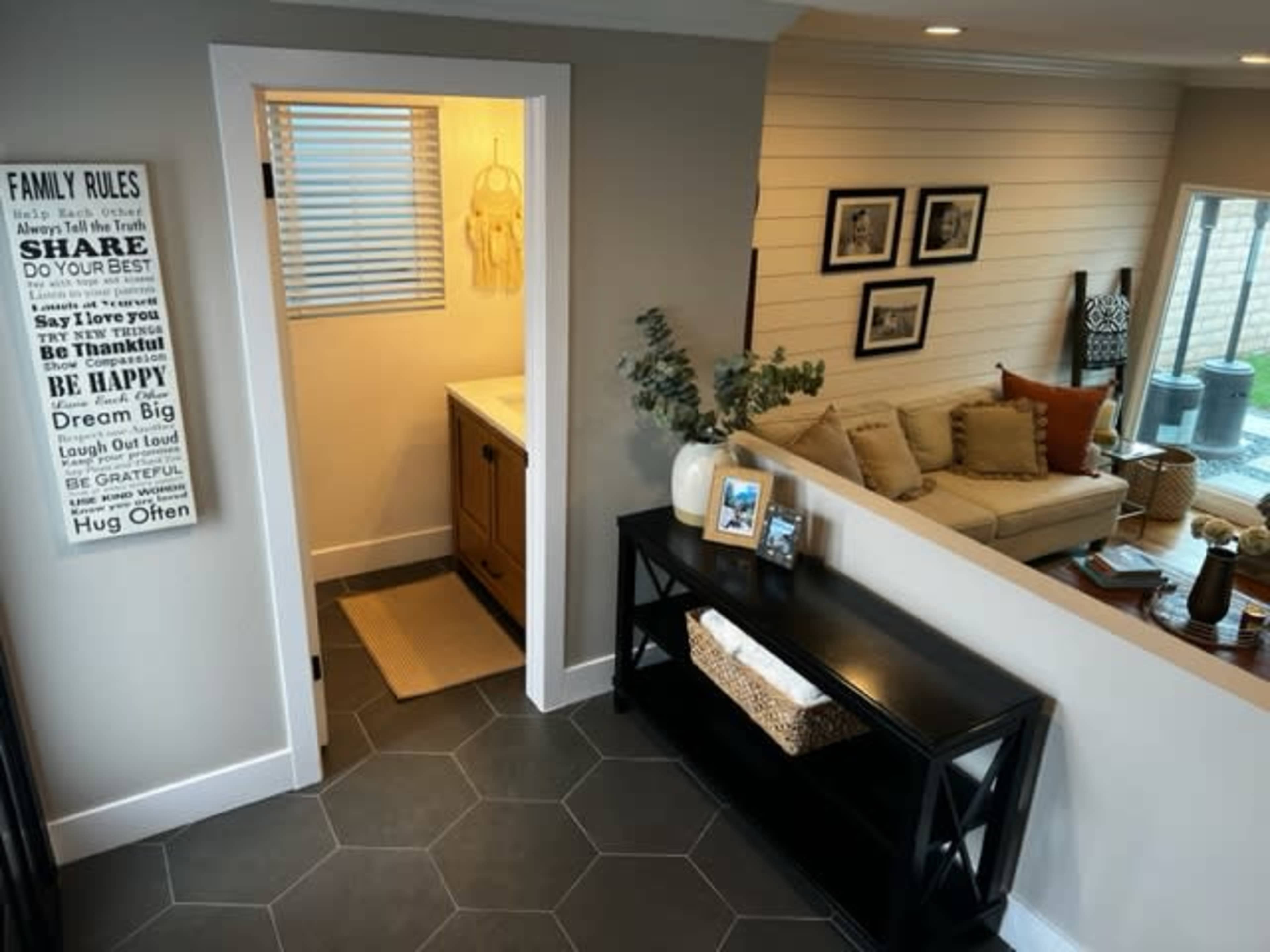 The image shows a modern entryway leading to a living area, with a console table, decorations, and a doorway that opens to a bathroom.