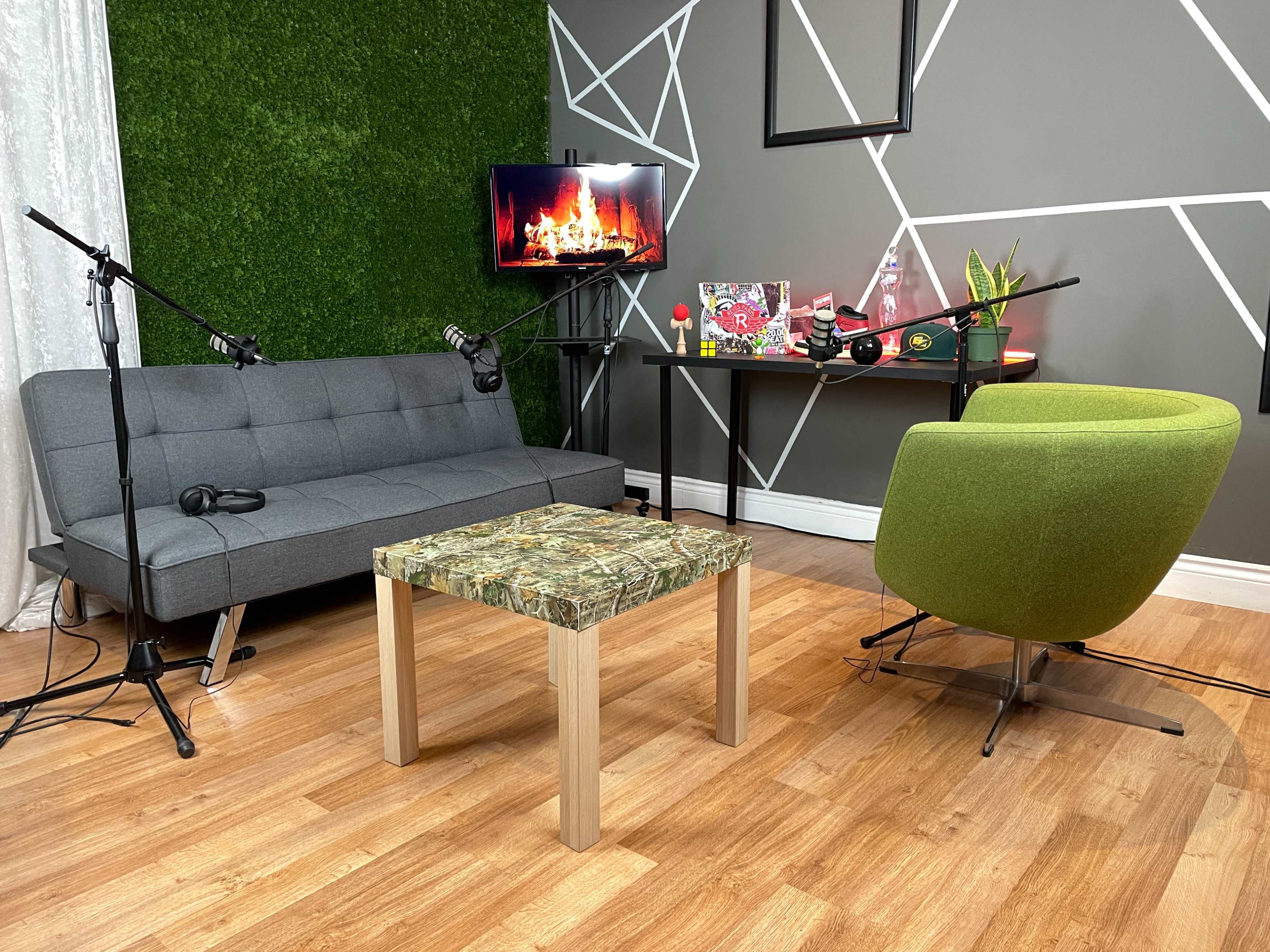 A cozy living room setup with a gray sofa, green chair, a wooden table with a camouflage design, and a wall-mounted TV displaying a fireplace scene.