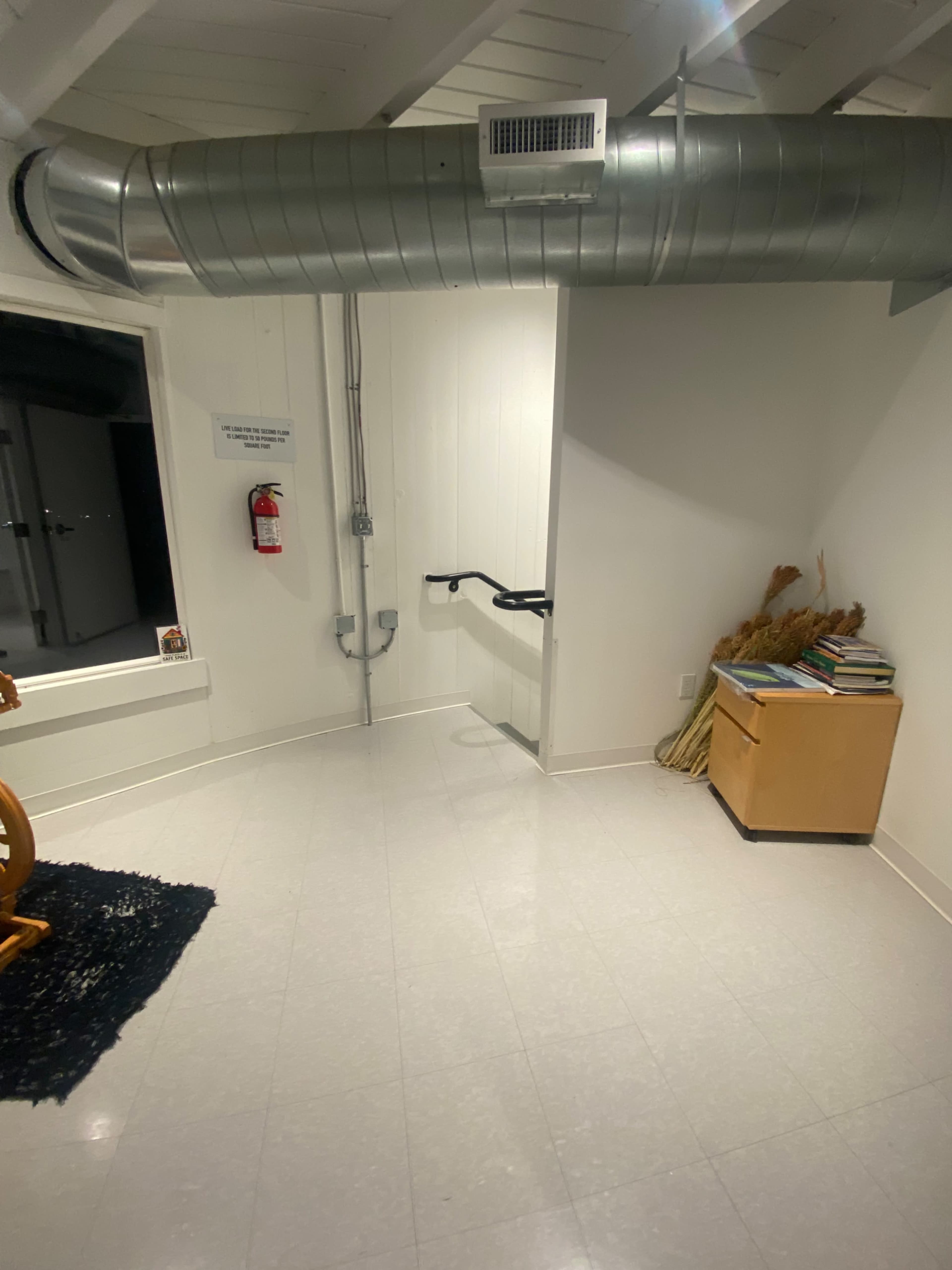 The image shows a dimly lit room with a staircase and handrail leading to another level, a metal duct overhead, and a small wooden cabinet with books next to a bundle of dried plants.