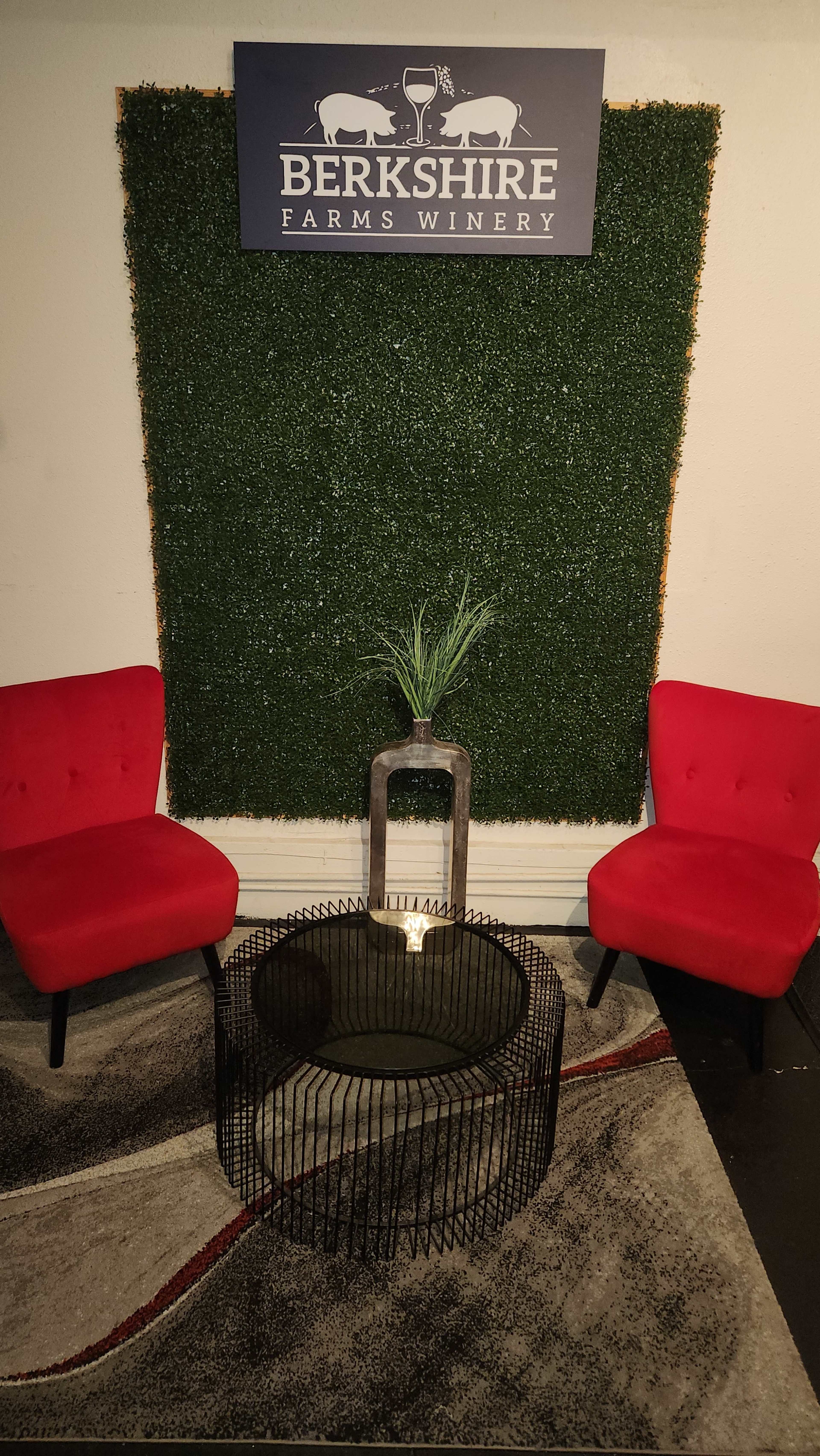 Two red chairs and a metal coffee table on a patterned rug, set against a greenery backdrop with a sign that reads "Berkshire Farms Winery."