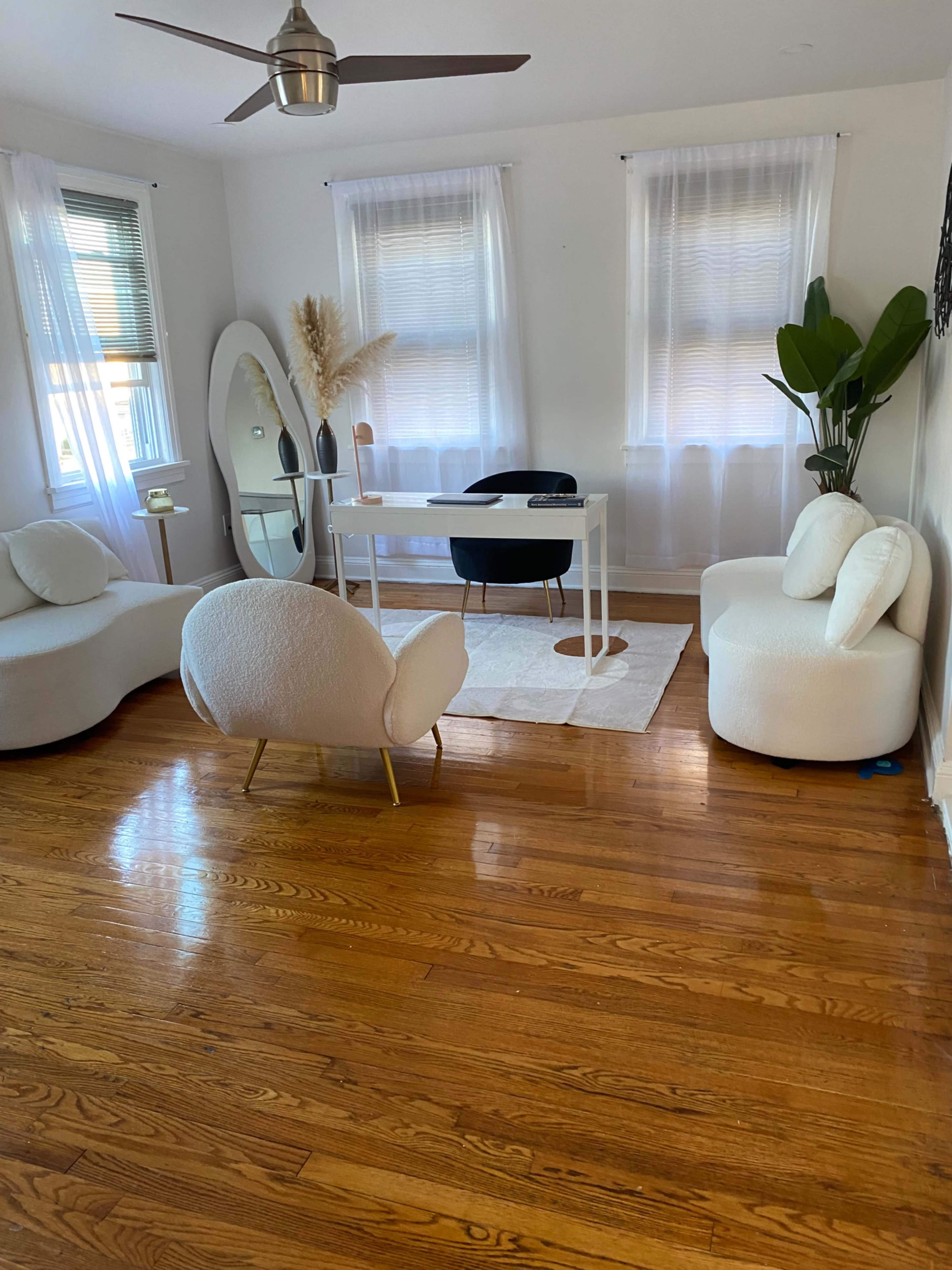 A well-lit room features a white desk and chairs, large windows with sheer curtains, and potted plants, all set on polished hardwood flooring.