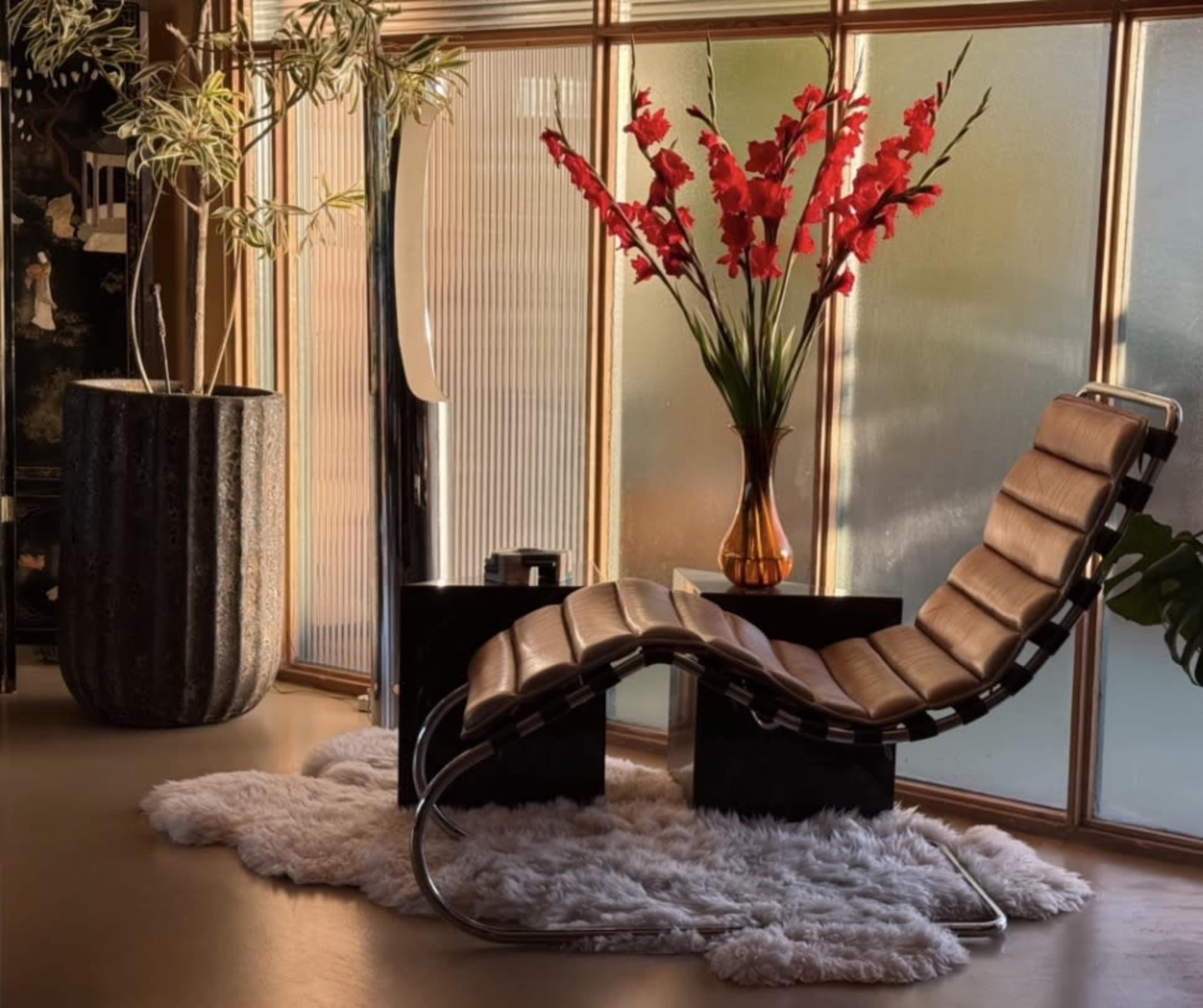 A lounge chair is positioned next to a black table, adorned with a vase of red flowers, all set against a backdrop of large windows.