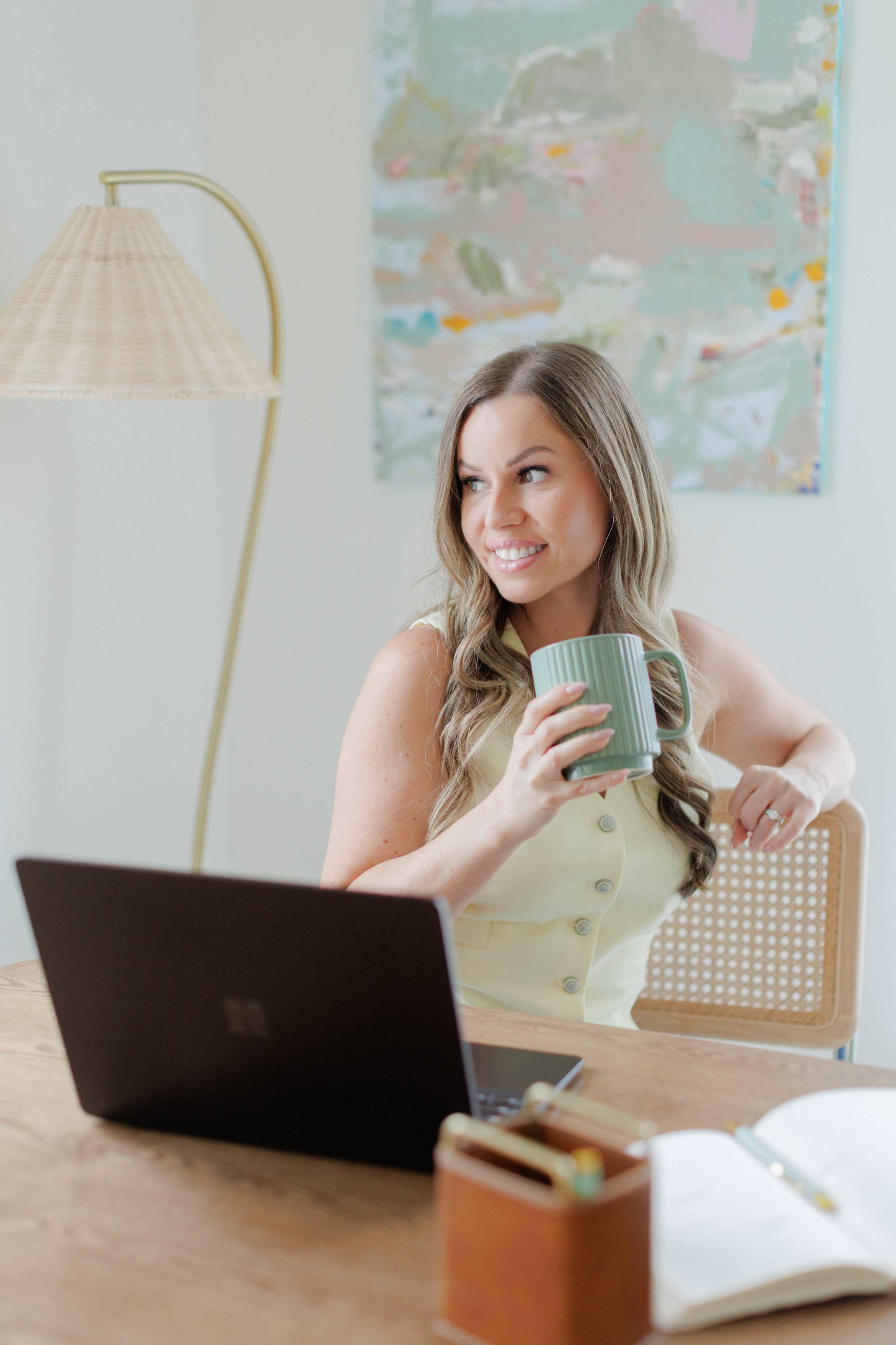 A woman sitting at a wooden desk holds a green mug while looking off to the side, with a laptop and notebook in front of her.