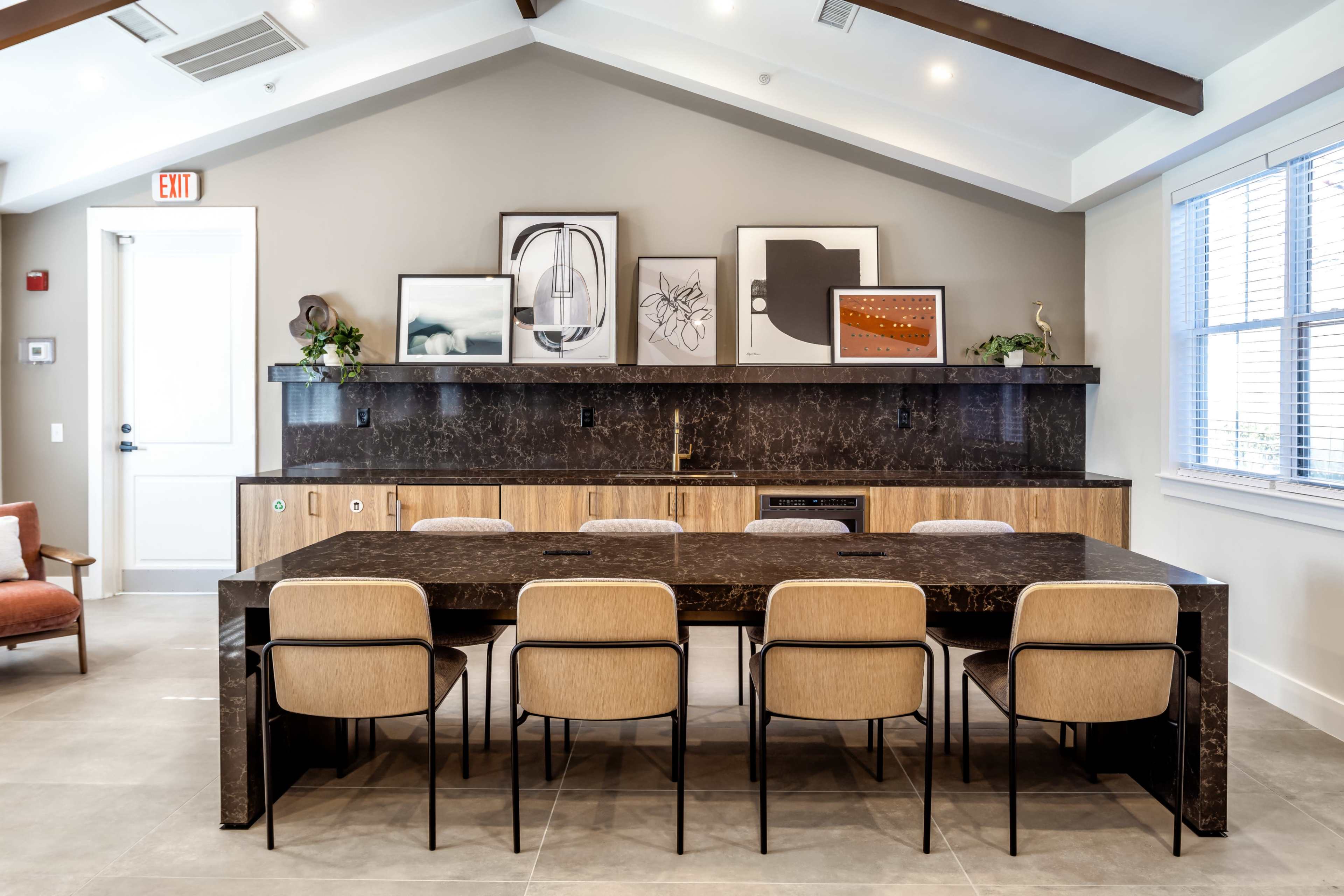 A modern dining area with a large black table surrounded by six beige chairs, set against a backdrop of wall art and a dark marble countertop.