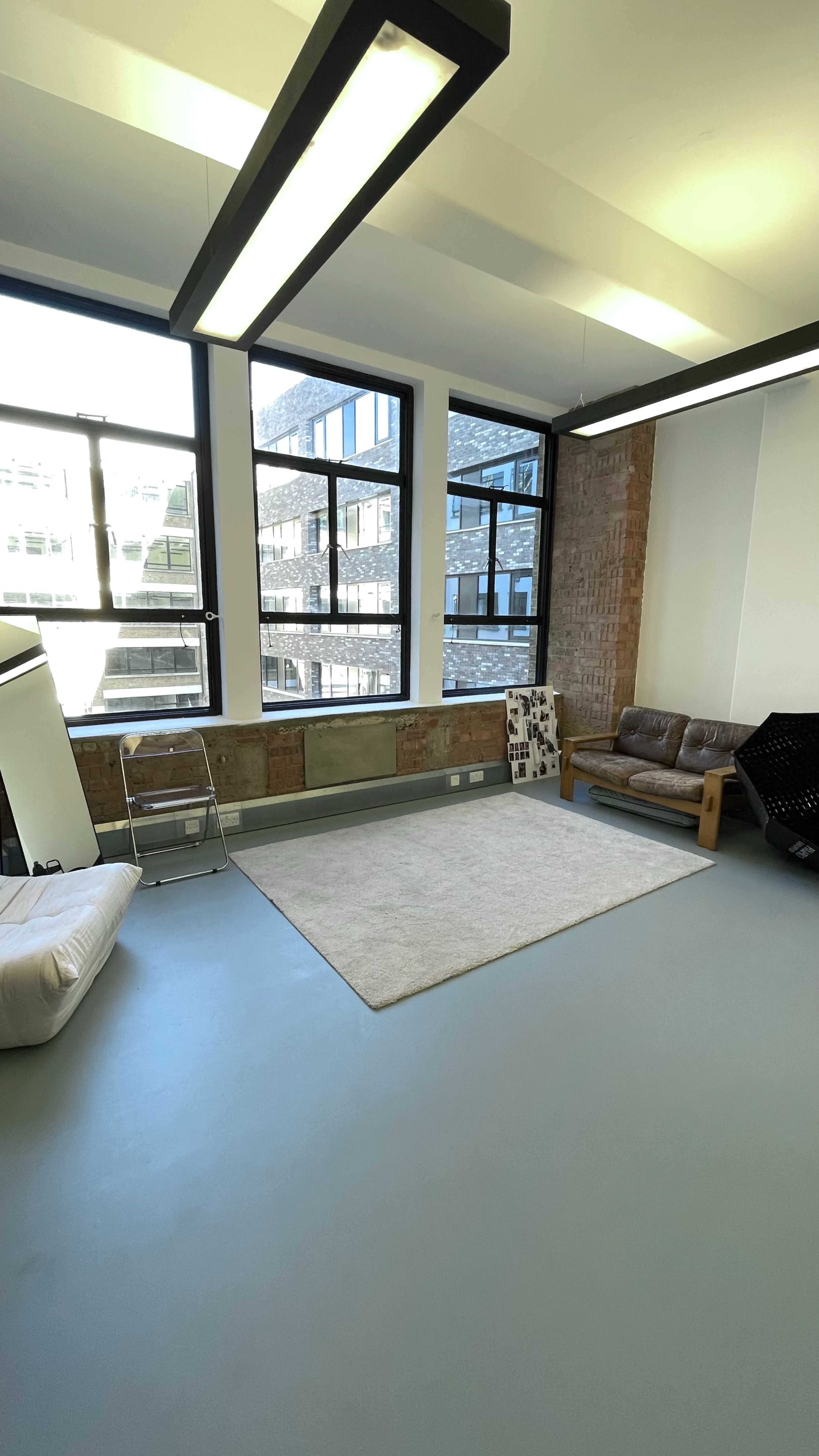 The image shows a spacious, minimalistic room with large windows, a couch, and a light-colored rug on a gray floor.