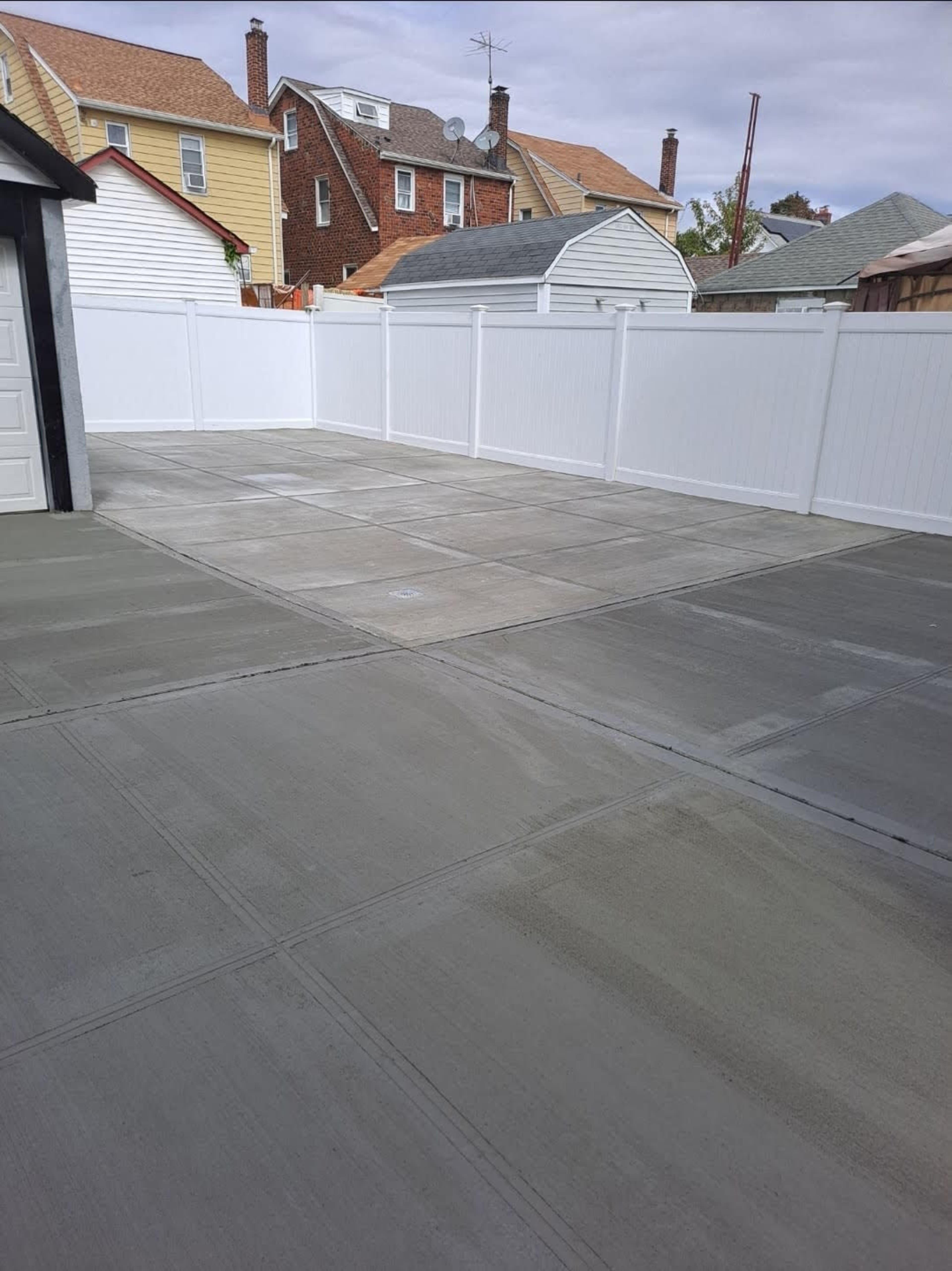 A newly poured concrete surface in a backyard, enclosed by a white fence, with several houses visible in the background.