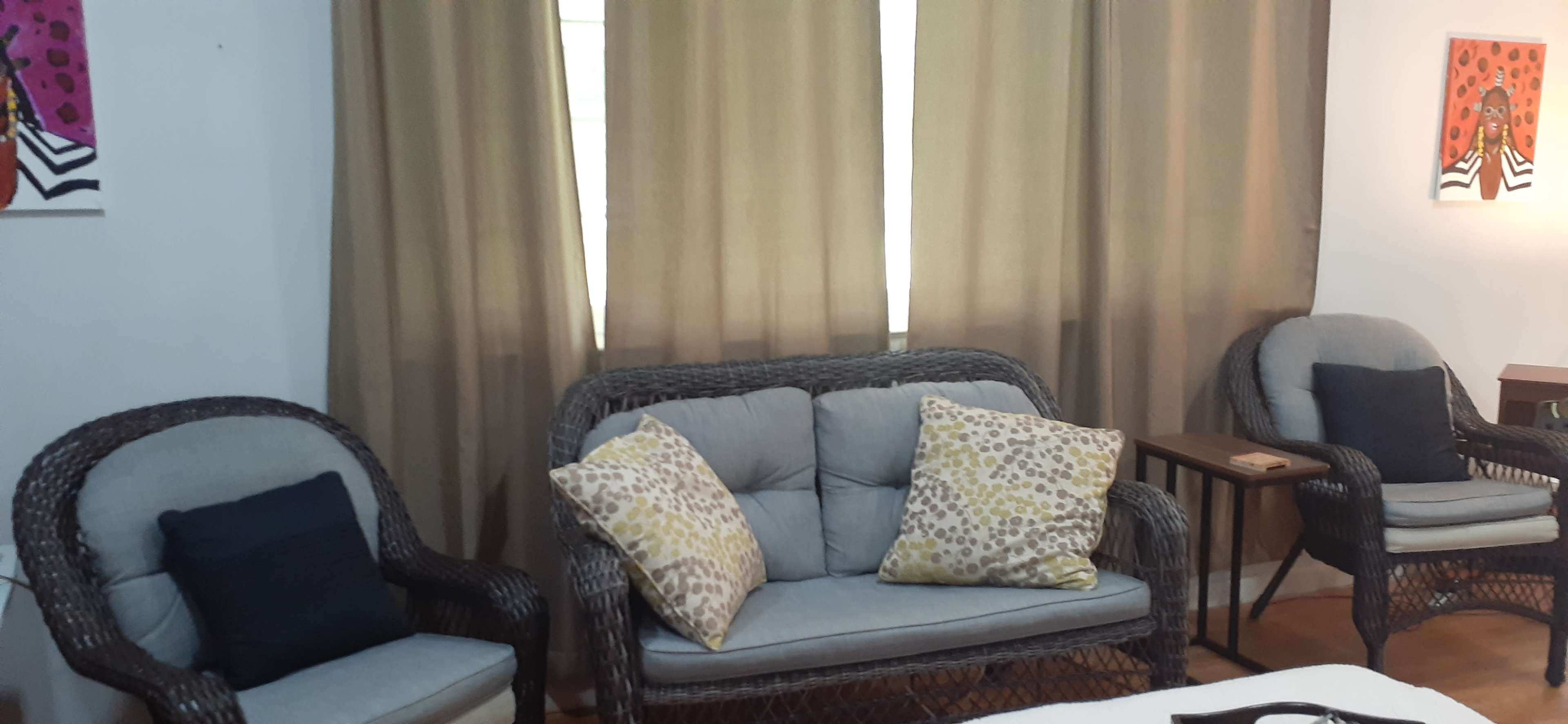 A seating area features a gray sofa with patterned cushions and two wicker chairs beside a small table, all set against a backdrop of curtains and wall art.
