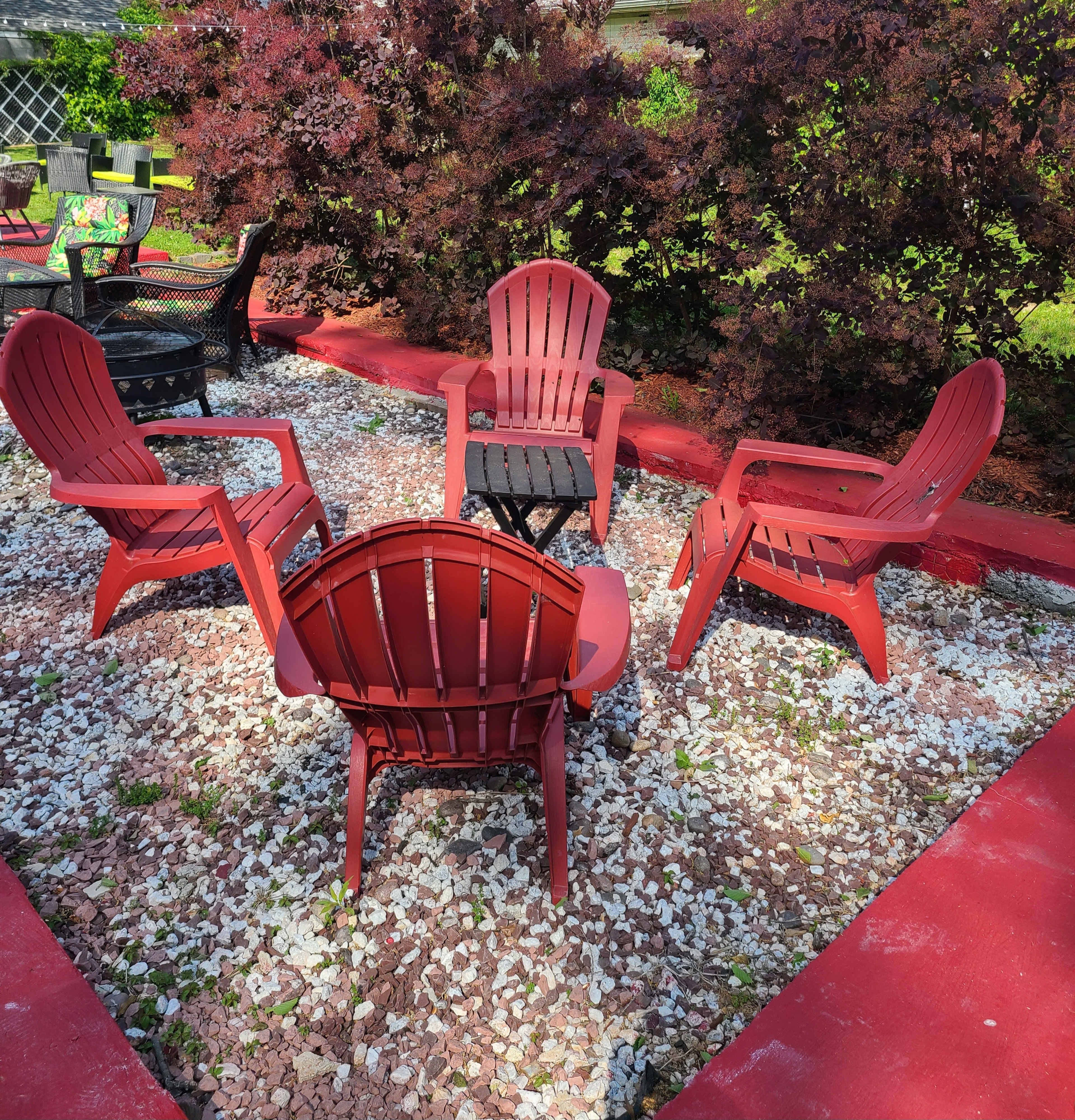 Four red Adirondack chairs are arranged in a circular formation around a small black table on a gravel surface, surrounded by landscaped bushes.