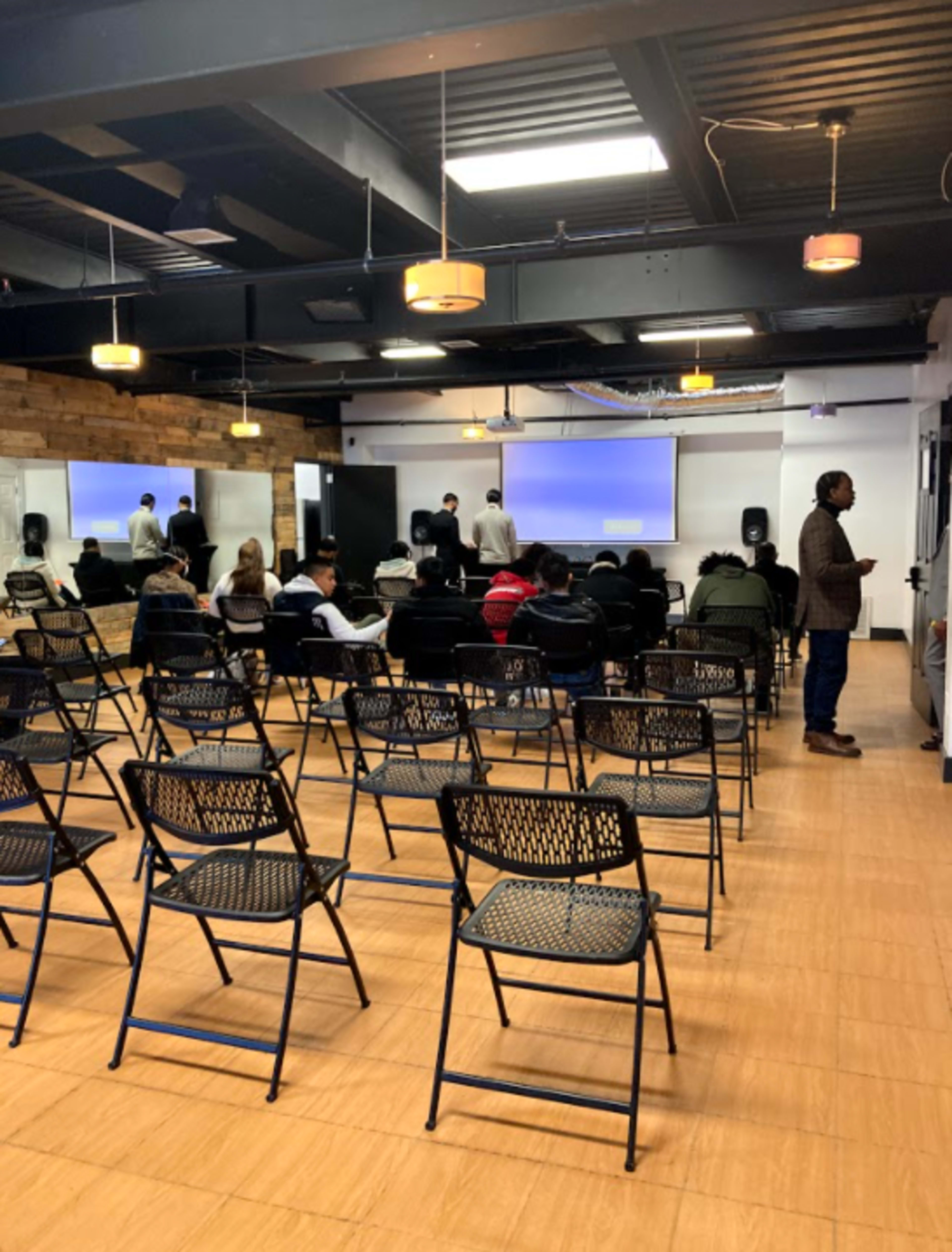 A crowded seminar room with rows of empty chairs facing a presentation screen, while a few individuals stand near the front.